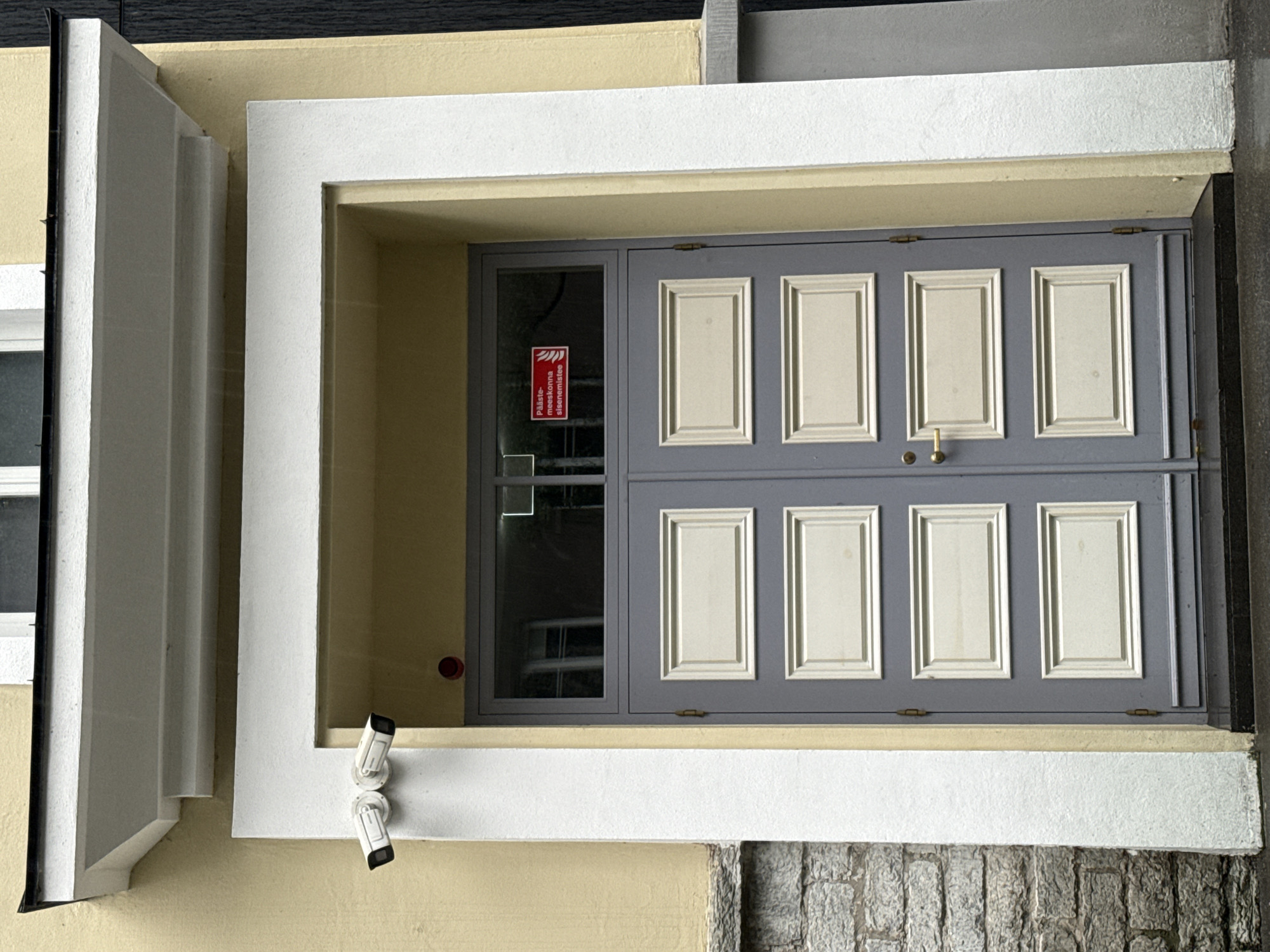 The image shows a gray double door with three panels on each side, each panel having a lighter beige inset. A gold doorknob is visible on the right door. Above the doors, there is a rectangular window with reflections of buildings and windows. On the glass of the upper part of the door, a red and white sign is affixed. Two white security cameras are mounted on the left side of the white archway framing the entrance. The building exterior is painted yellow with a gray stone foundation on the left and a dark vertical element on the right. The scene appears to be an entrance to a building in Tallinn, Estonia, as suggested by the Estonian text on the sign which translates to "Fire department interior access only." The ground in front of the door is wet, indicating it may have been raining.