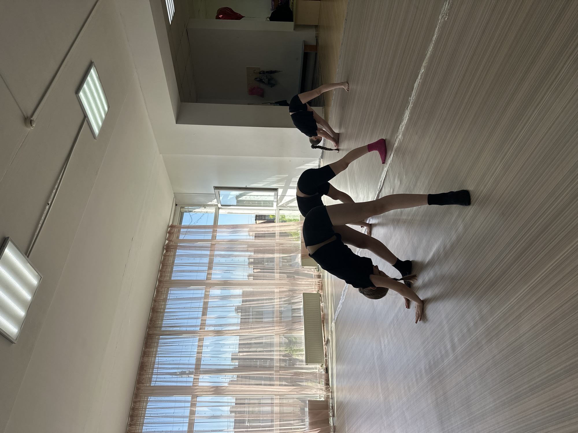 Two people are practicing gymnastics in a well-lit dance studio. Both are bent over in a backbend position. They are wearing black athletic clothing. The floor is light-colored wood. Tall windows are visible in the background. This photo was taken in Votkinsk, Russia.