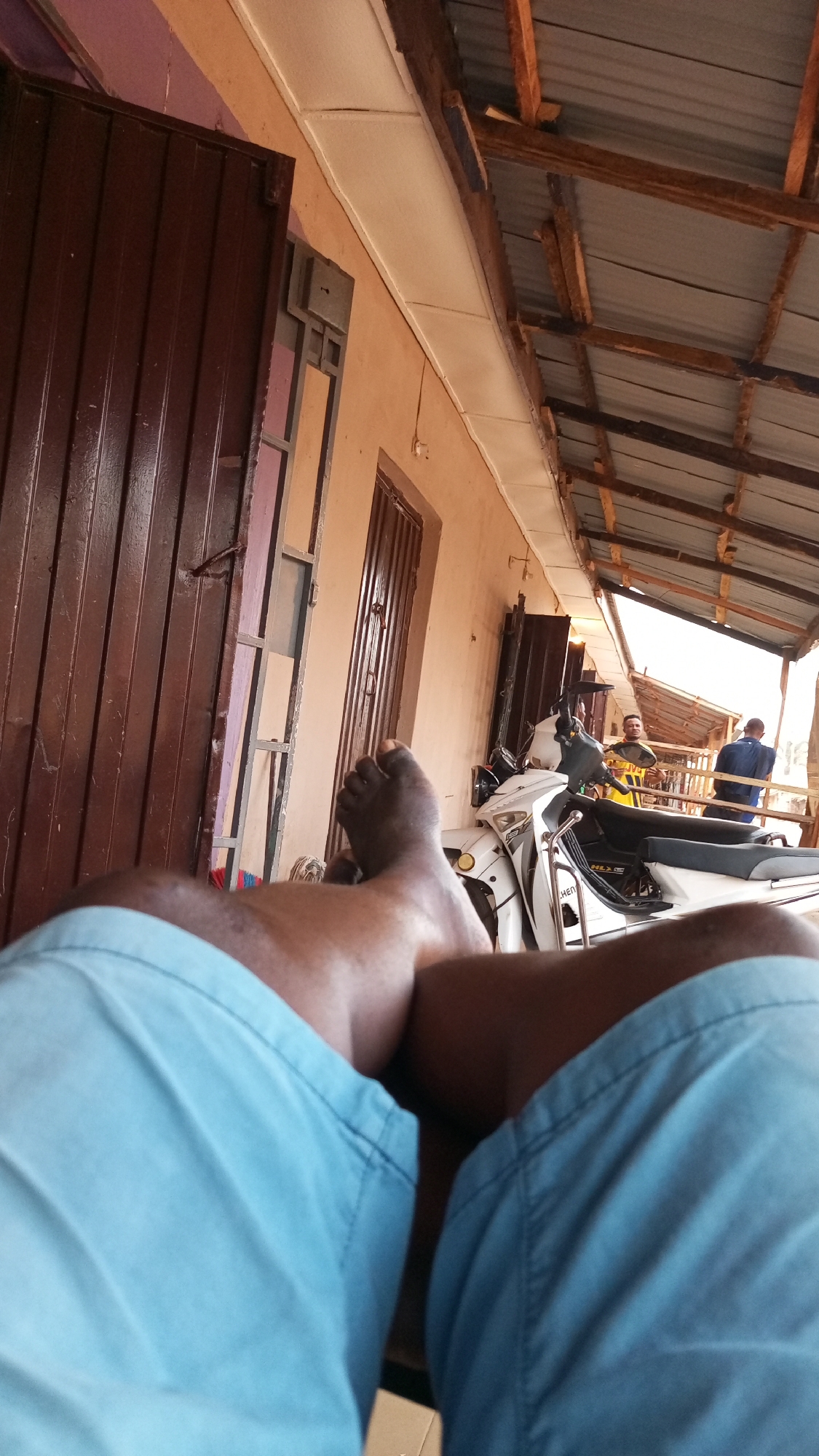 The image shows a first-person view of a person sitting outdoors, likely on a porch or covered area, in Sabon Tasha, Nigeria. The person is wearing light blue shorts and their bare feet are visible. A white scooter or small motorcycle is parked nearby. In the background, two people are visible, one wearing a yellow shirt and the other a blue shirt. The scene is in front of a building with dark doors and windows. The structure overhead has a corrugated metal roof supported by wooden beams.