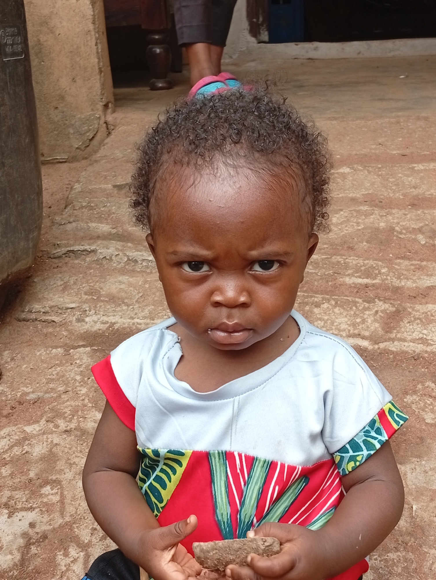 A young child with dark skin and curly hair sits in front of a dirt or concrete ground. The child wears a shirt with a red, yellow, and green pattern. The child's left hand holds a small, brown object, possibly a piece of wood or root. The background is out of focus, but appears to be an outdoor setting with a wall and some furniture. The overall scene is a candid portrait of the child. The image does not contain enough information to determine the specific location within Adda, Nigeria.