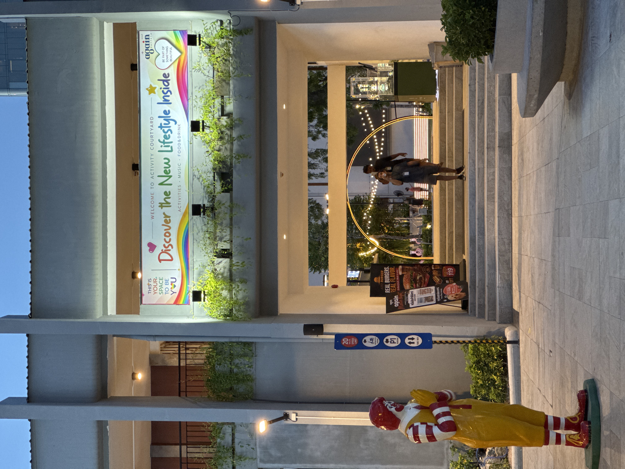The image depicts an outdoor scene in Pattaya City, Thailand, at what appears to be an entertainment or retail complex. In the foreground, a statue of Ronald McDonald stands to the left, facing right with his hands pressed together in a prayer-like gesture. He is dressed in his iconic red and yellow attire. To the right of Ronald McDonald, a blue signpost displays information, including a speed limit of 20 km/h, a motorcycle icon, a parking symbol labeled "P1," and a "TWO WAY" indicator. Further to the right, a display board advertises "REAL BURGERS. REAL FLAVOR." from McDonald's.
The background reveals an entranceway leading into a courtyard. Above the entrance, a banner reads "WELCOME TO ACTIVITY COURTYARD" and prominently features the text "Discover the New Lifestyle Inside" with a rainbow graphic. The McDonald's logo and the words "again PATTAYA" are also visible on the banner. Within the courtyard, a golden arch adorned with string lights creates a focal point, with several people visible walking through it. The environment is softly lit by artificial lights, suggesting it is either dusk or evening. Greenery, in the form of hanging plants and bushes, is integrated into the architecture, adding a natural element to the urban setting.