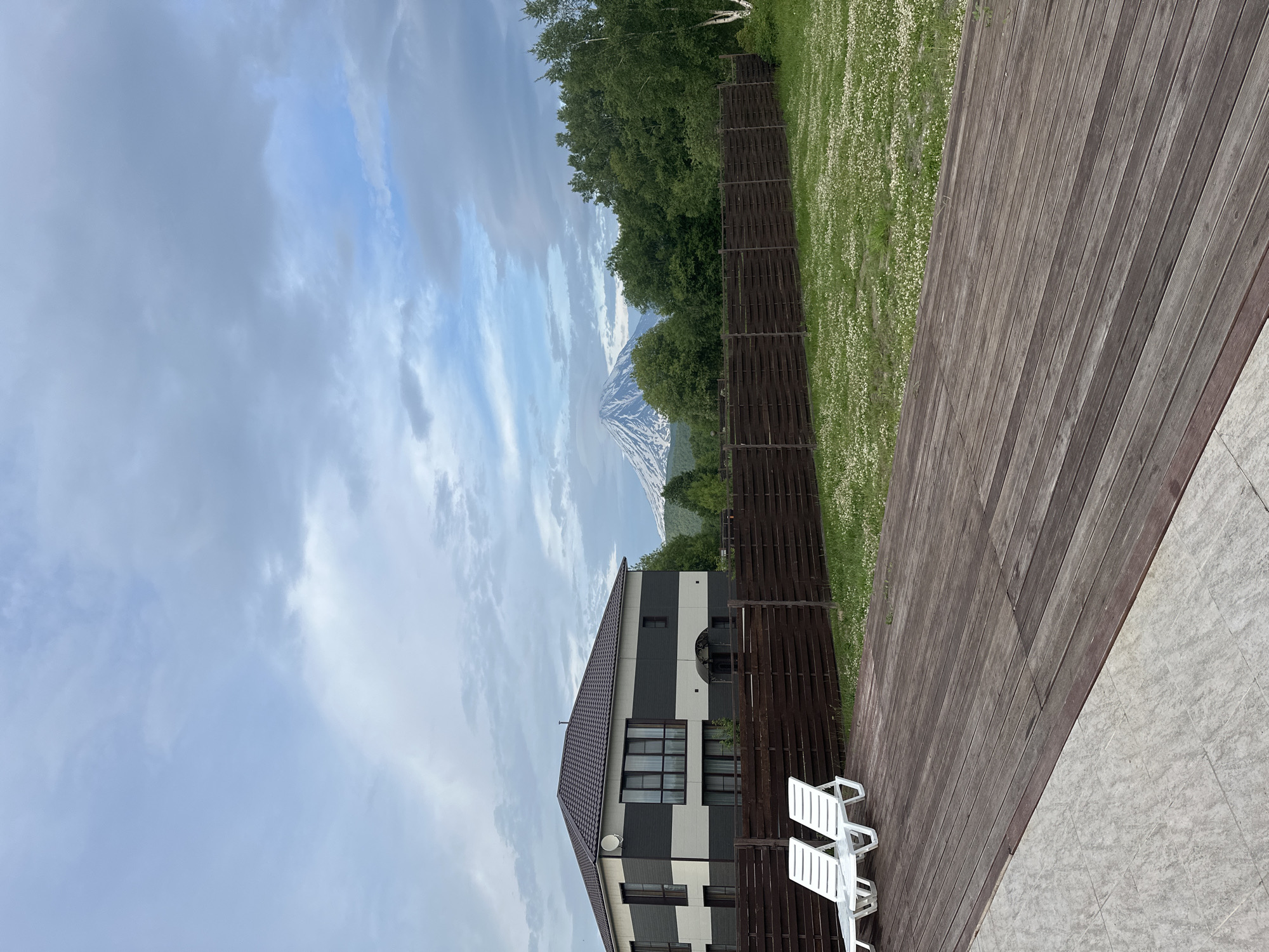 The image depicts a modern two-story house with a dark brown, pitched roof, featuring horizontal bands of dark grey and off-white on its exterior. The house has numerous windows, some tinted, and a satellite dish mounted on the upper facade. In front of the house, a dark brown wooden fence encloses a grassy yard dotted with small white flowers. Two white lounge chairs are positioned on a dark wooden deck in the foreground. Beyond the fence and the house, a majestic, snow-capped mountain, possibly a volcano, dominates the background under a cloudy sky. Lush green trees surround the mountain and extend to the right side of the frame. The scene appears to be set in a semi-rural or suburban area, likely in Yuzhnyye Koryaki, Russia, given the prominent volcanic landscape. There are no people or animals visible in the image.
