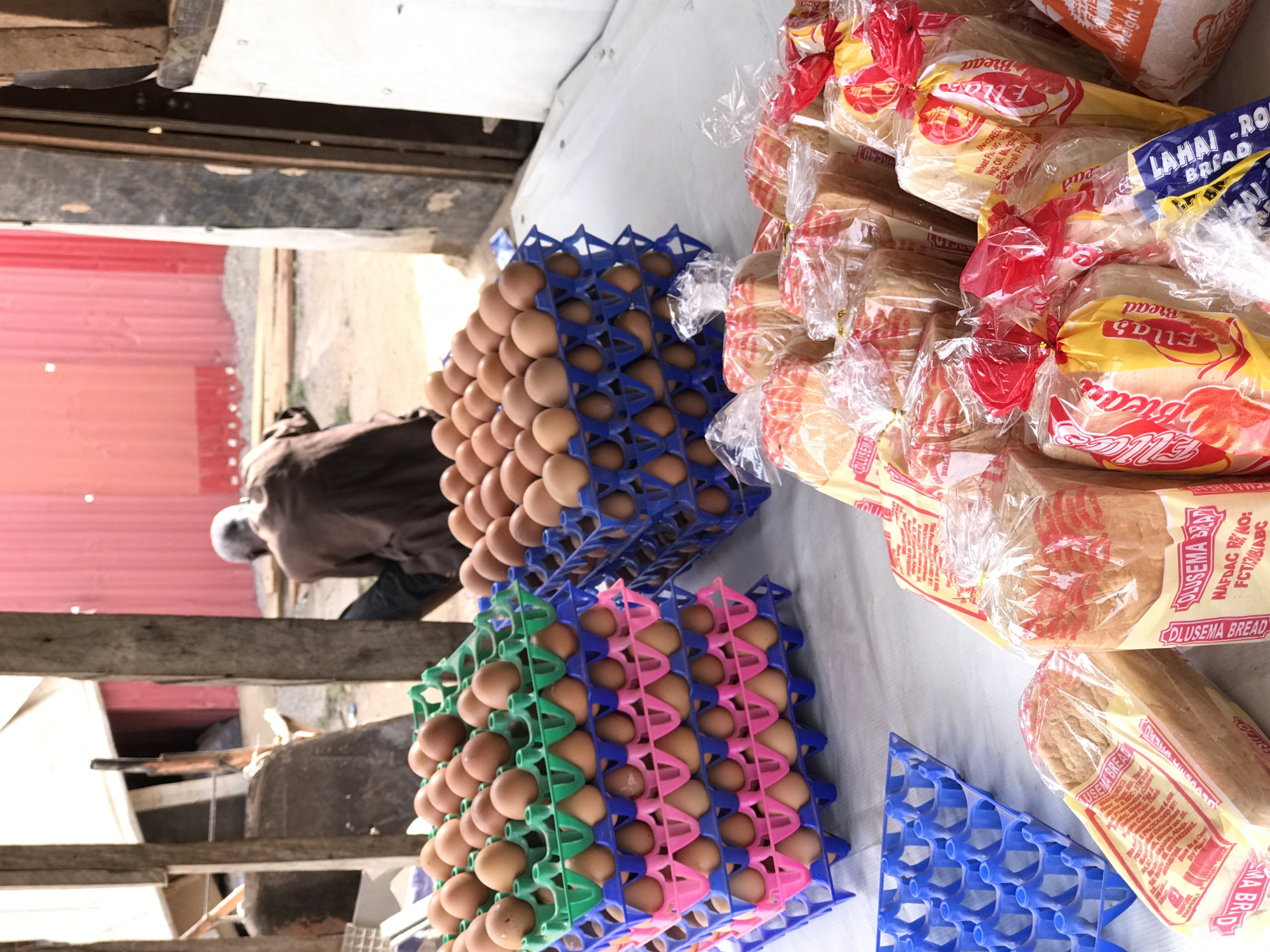 The image depicts a market stall or shop, likely in Abuja, Nigeria, showcasing everyday food items. The primary subjects are numerous trays of eggs and stacks of packaged bread loaves, displayed on a light-colored surface, possibly a table or tarp.
Multiple plastic trays, in blue, green, and pink colors, are filled with brown chicken eggs. Some empty blue egg trays are also visible. Next to the eggs, dozens of loaves of bread are individually wrapped in clear plastic bags, predominantly with red and yellow labels, and some with blue labels.
Visible text on the bread packaging includes "DLUSEMA BREAD" (which appears frequently), "LAHAI-RO BREAD," and "Ellas Bread." Further details like "INGREDIENTS: Wheat Flour, Veg. Oil, Margarine, Edible Salt, Royal) Flour," "NAFDAC RE NO: FCT/3/8(ABC," and "Store in a Cool Dry Place" are legible from the OCR.
In the background, a person, possibly a vendor, is hunched over. They wear a dark top and a light head covering, with their back mostly turned to the viewer. The background features a vibrant red corrugated metal wall and various wooden beams and structures, suggesting an outdoor or semi-outdoor market environment. The scene is brightly lit by natural light, indicating it is daytime. The overall impression is that of a bustling local commerce setting.