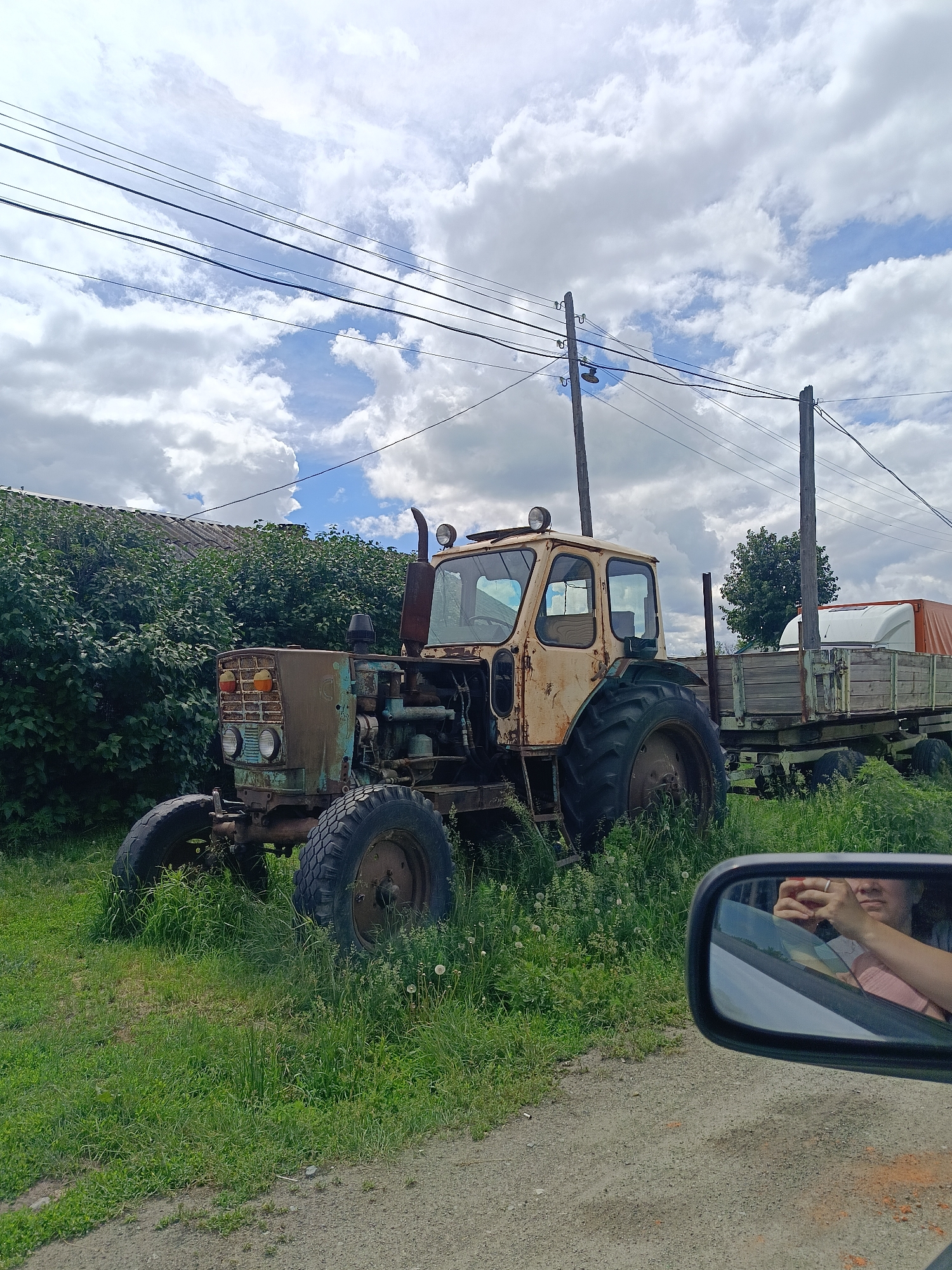 An abandoned tractor, painted beige and teal, sits in tall green grass beside a wooden trailer filled with cargo covered by an orange tarp. The tractor is weathered and rusty, with large, treaded tires. Behind the tractor, a dense line of green foliage borders a building with a grey, corrugated roof. Overhead, power lines stretch across a partly cloudy sky with fluffy white clouds. In the foreground, the reflection in a car's side mirror shows a person wearing a pink shirt and holding a phone. The scene is set on a dirt road in the city of Kamennoye, Russia.