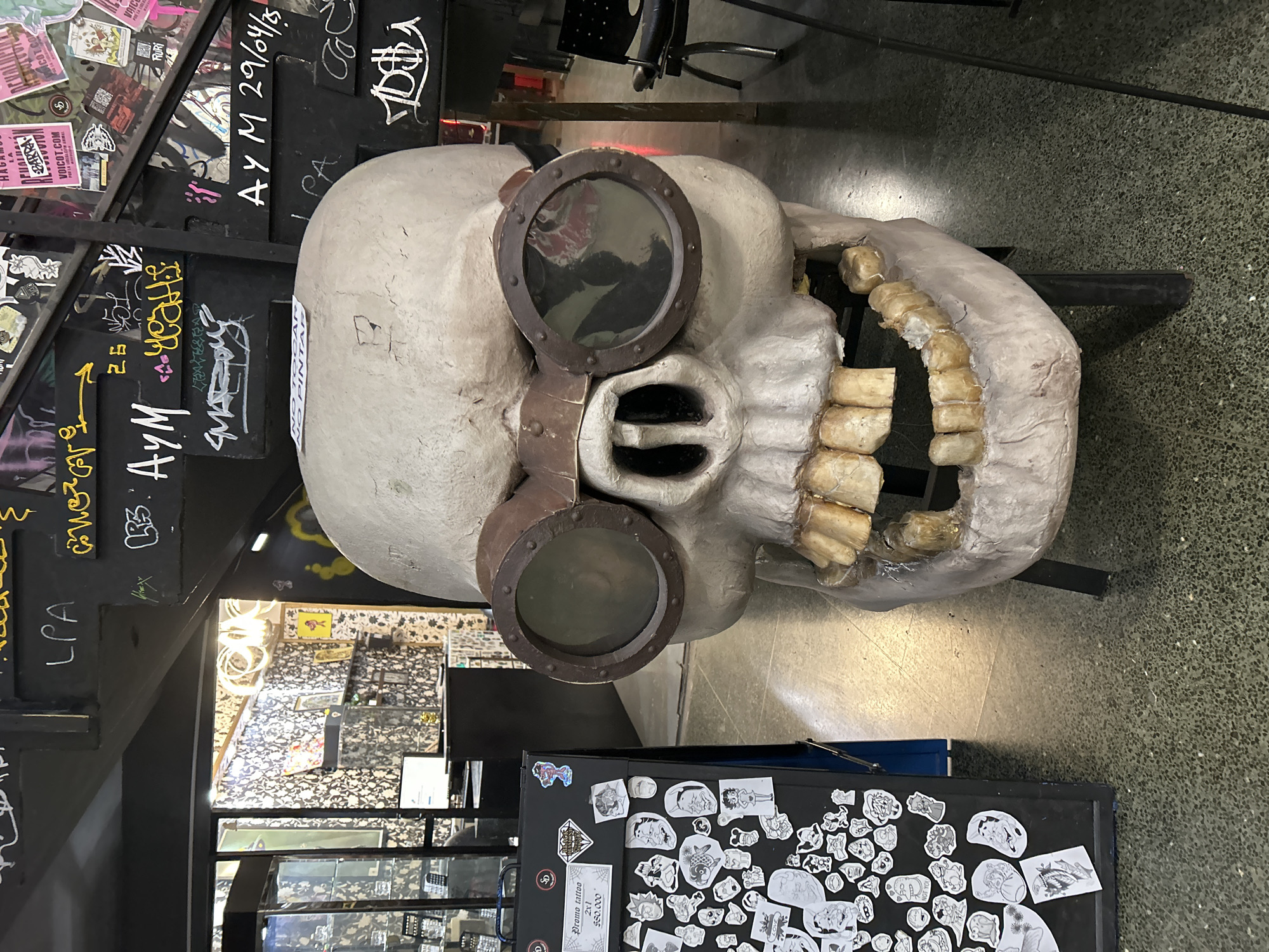 An interior shot captures a large, stylized, off-white skull wearing dark, riveted goggles, positioned as a prominent display item. The skull features chunky, off-white teeth in an open jaw and has a "NO PINTAR" (Do Not Paint) sticker on its forehead.
The setting appears to be an urban or alternative venue, possibly a tattoo shop or bar, in Buenos Aires, Argentina. The background features dark walls heavily covered in graffiti tags and various stickers, contributing to a vibrant, counter-cultural aesthetic. Some of the graffiti includes "AYM 29/04/5", "LPA", "yeah", and "VOICOT.COM".
In the lower right foreground, a dark cabinet or table is completely adorned with numerous small, black-and-white tattoo flash designs, many depicting cartoon characters or quirky figures. A sign on this table advertises a "Promo tattoo 2x1 $80.000". The floor is a speckled, dark grey concrete or terrazzo. The overall ambiance suggests a creative, edgy, and informal space. There is no explicit human activity, but the arrangement implies a public or commercial establishment.
