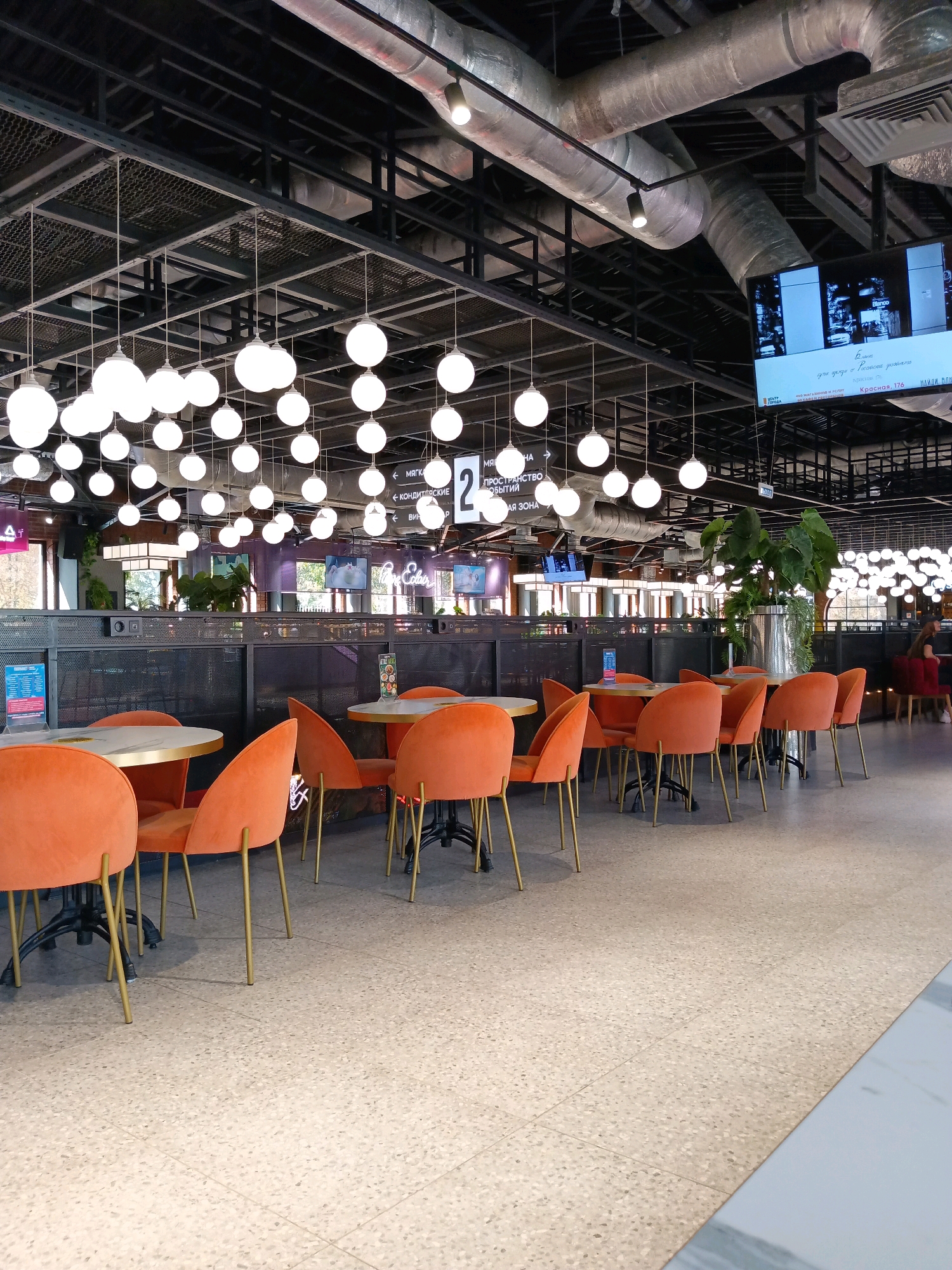 The image shows a modern food court area with multiple tables and chairs, located in Krasnodar, Russia. The tables are round with marble-like tops and gold-colored legs, and the chairs are upholstered in a vibrant orange velvet. Overhead, a complex network of metal beams supports numerous round, white pendant lights. Large industrial ventilation ducts are also visible on the ceiling. In the background, there are large screens displaying content, and a large potted plant with broad leaves adds a touch of greenery. The floor is tiled with a light-colored terrazzo pattern. Several signs with Russian text are visible, including one indicating "MEAT" and another with the number "2". The overall impression is of a stylish and contemporary dining or gathering space.