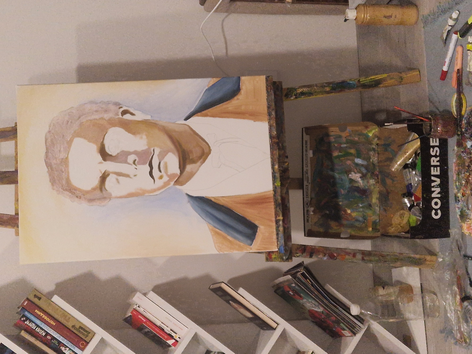 A portrait painting on an easel is the central focus of the image. The unfinished portrait depicts a man, mostly in shades of brown and white, with some blue visible around the collar area. The canvas is held by a wooden easel that appears to be splattered with various colors of paint. To the right of the painting, there's a disorganized collection of art supplies including a Converse-branded box. Further to the right, a collection of various art-related materials is spread across a surface. Below the canvas, there is a shelf with books and other items. The books appear to be a mix of fiction and non-fiction. The background is a plain, light-colored wall.