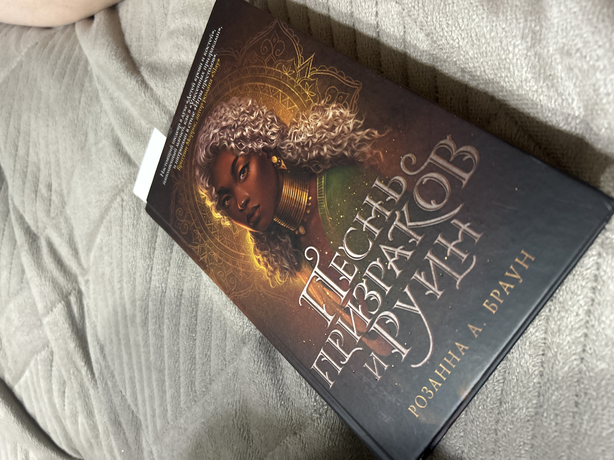 The image shows a book with a dark cover, likely on a textured, light gray surface. The book cover features an illustration of a woman with dark skin, curly hair, and golden jewelry against a background with a stylized golden pattern. The title is written in a decorative Cyrillic script, "Песнь призраков и руин," and the author's name, "Розанна А. Браун," is below the title. A white bookmark or piece of paper is sticking out from the top of the book. The book is lying on the surface at an angle.
There is no information in the image to connect it to Chaikovsky, Russia.