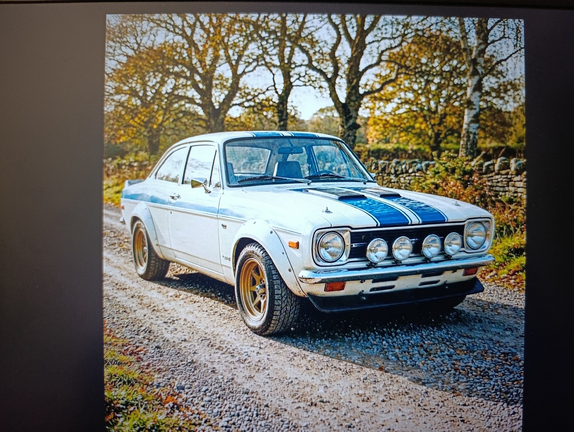 A white Ford Escort Mk2 is parked on a gravel road. The car has blue racing stripes and gold wheels. There are four fog lights mounted on the front bumper. Trees are visible in the background, with leaves in autumn colors. A stone wall runs along the right side of the road behind the vehicle. The location is likely rural, as there are no visible buildings or signs of urban development. It is difficult to verify that this is Іргелі ауылдық округі, Kazakhstan. There are no people visible in the image.