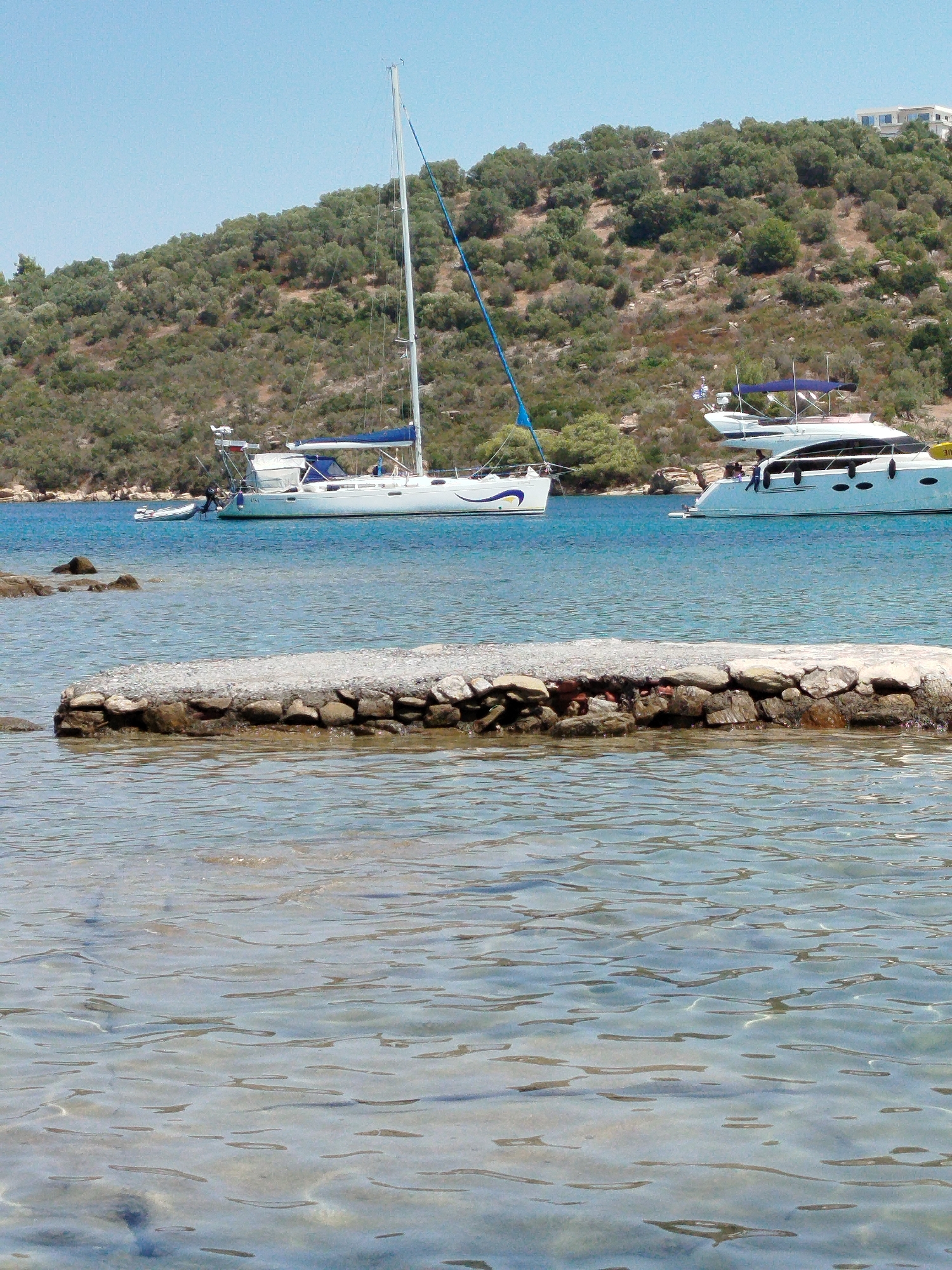 A sunlit, daytime, medium-shot captures a tranquil coastal scene with two yachts anchored in clear, blue water. In the foreground, a low, rough stone jetty extends into the water, its surface uneven and mottled. The water is shallow, allowing the sandy bottom to be partially visible beneath the ripples. In the mid-ground, a white sailboat with a blue canopy and a single mast is moored, its hull adorned with a yellow and blue graphic. To its right, a larger, sleeker white motor yacht with multiple windows and a dark blue trim is also anchored. Beyond the water, a verdant, hilly landscape rises, densely covered with olive trees and scattered rocks, under a clear, pale blue sky. A few white buildings are visible atop the hill in the distance. The overall impression is one of peaceful summer leisure in a Mediterranean setting.