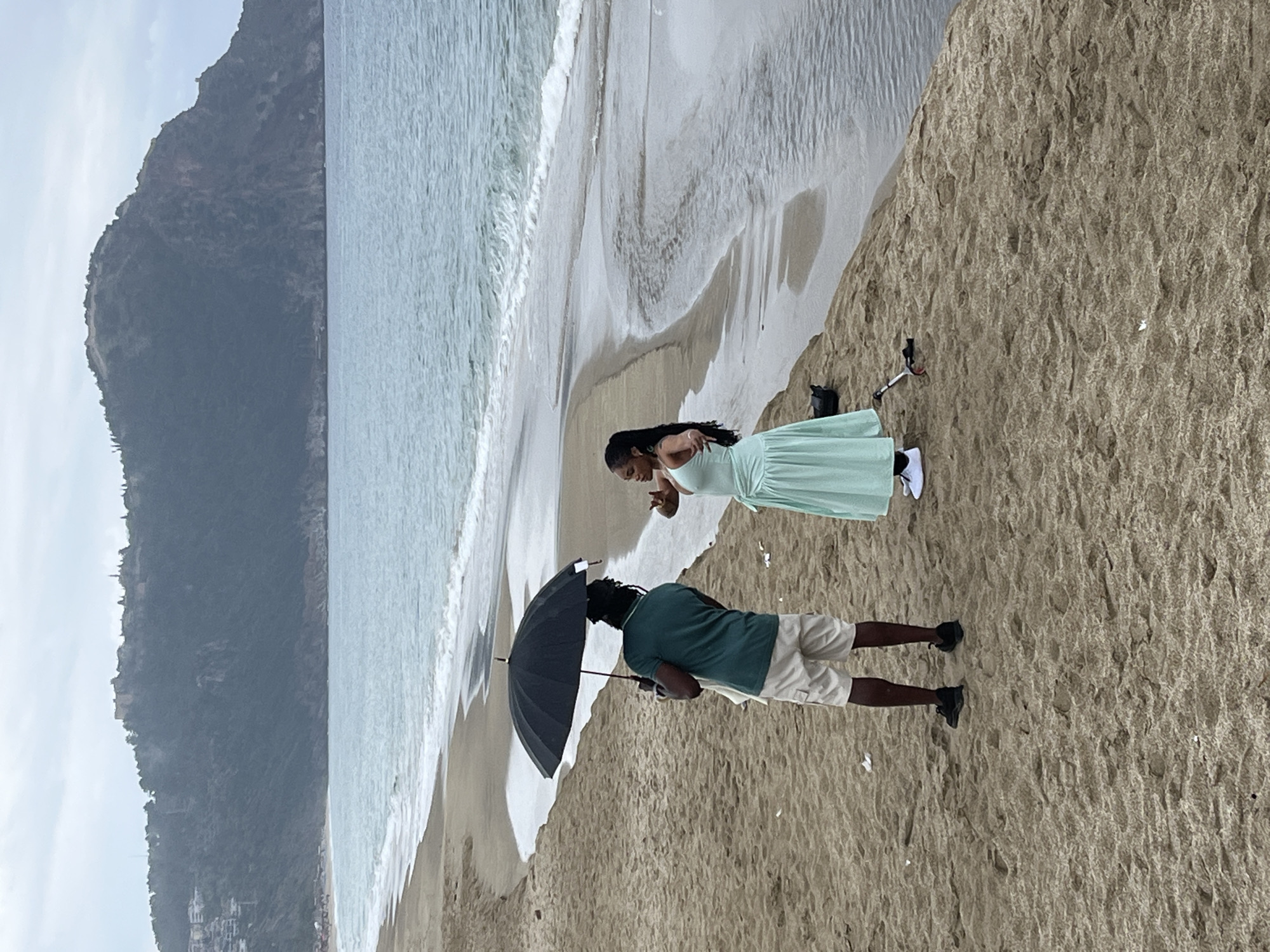 A medium-range shot captures a man and a woman on a sandy beach with an umbrella. The woman, wearing a light green dress, is turned toward the man. He is holding a black umbrella. They are standing on the beach near the water's edge. The man is wearing a green shirt and khaki shorts. A mountain rises in the background along the coast.
Based on the visible mountain range and beach, the location is Alanya, Turkey.