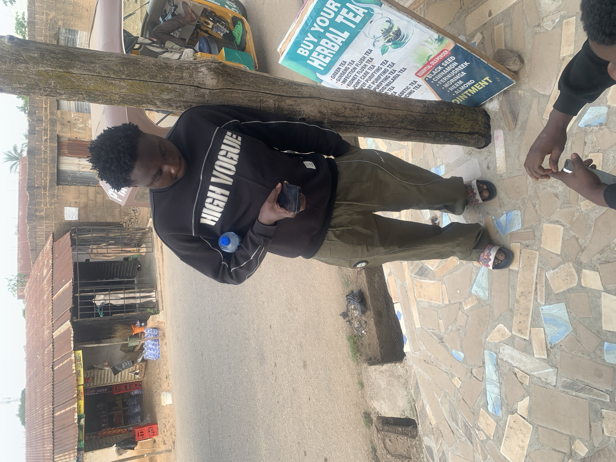 This daytime image, set in Jos, Nigeria, features a young man centrally positioned, leaning against a rough wooden pole. He wears a black sweatshirt with "HIGH VOGUE" emblazoned on it, olive green cargo pants, and patterned sandals. In his left hand, he holds a dark smartphone, and a clear plastic water bottle is visible near his right arm. He looks downwards, appearing contemplative.
To the right, the hands of another person are visible, also holding and interacting with a smartphone. The ground in the foreground is a mosaic of tan, white, and blue-gray tiles, transitioning to a paved road further back.
Behind the main subject, a vibrant sign advertises "BUY YOUR HERBAL TEA" and "ESSENTIAL OILS POWDER / OINTMENT." Visible tea varieties include Green Tea, Ginseng Tea, Infection Flush Tea, Kidney Flush Tea, Joint Care Tea, Liver Purifying Tea, Heart Purifying Tea, Malaria Tea, and Diabetic Tea. The essential oils, powders, and ointment ingredients listed are Black Seed, Cinnamon, Fenugreek, Moringa, Neem, and Almond.
In the background, a yellow and beige tricycle (Keke Napep) is partially visible. Further along the street, several single-story buildings with corrugated iron roofs line the road, suggesting a commercial or residential area. The scene depicts a casual street setting under bright sunlight.