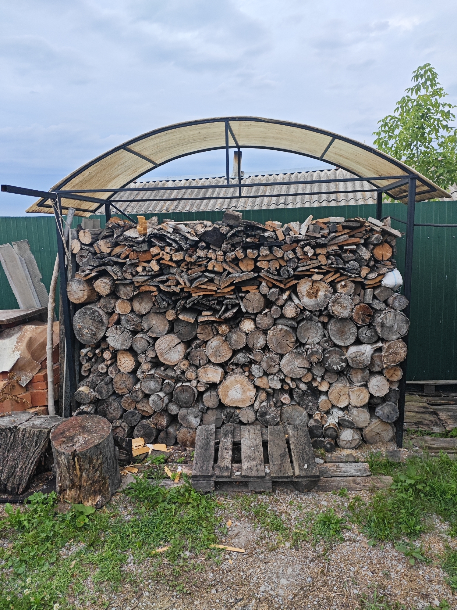 The image depicts a woodpile stored under a metal-framed awning with a translucent fabric roof. The woodpile is substantial, composed of various sizes of logs and split wood, neatly stacked against a green corrugated metal fence. The logs are cut to different lengths and diameters, with visible grain patterns and bark textures. Some of the wood appears to be seasoned. In front of the woodpile, there are two large tree stumps, one with prominent bark texture and the other with a smooth, cut surface. Partially visible to the left of the woodpile are some construction materials, including bricks and flat concrete-like slabs. The ground in front of the woodpile is covered with gravel and patches of green grass and weeds. The sky above is overcast with a cloudy, pale blue hue. The overall scene suggests an outdoor storage area, possibly in a rural or semi-rural setting. There are no people visible in the image. The city of Песчанка, Russia, is not discernible from the visual information provided in the image.