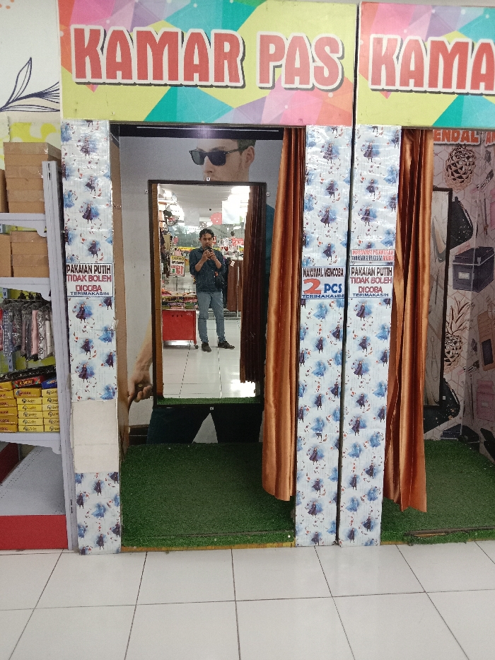 This image depicts the entrance to a fitting room area within a retail establishment in Langenharjo, Indonesia. The central feature is a doorway framed by two vertical panels adorned with a "Frozen" character pattern, specifically Elsa, Anna, and Olaf, set against a light blue and white background. Overhead, a large sign with a colorful geometric background reads "KAMAR PAS KAMAR" in prominent red letters.
Inside the fitting room area, which has an artificial green grass floor, a male individual is visible in the reflection of a large, black-framed rectangular mirror. He is standing, wearing a dark long-sleeved top or jacket, light-colored pants, and dark shoes, holding a black smartphone to take a picture of his reflection. Behind him, reflected in the mirror, is a portion of the store interior, including racks of clothing, a red display unit, and an overhead promotional image of a man wearing sunglasses.
Flanking the fitting room entrance are two brown/gold curtains, partially drawn. On the vertical patterned panels, two white signs with red text provide instructions:
1. The sign on the left reads: "PAKAIAN PUTIH TIDAK BOLEH DICCOBA TERIMAKASIH" (White clothes are not allowed to be tried on, thank you).
2. The sign on the right reads: "MAXIMAL MENCOBA 2 PCS TERIMAKASIH" (Maximum try on 2 pieces, thank you), with the "PAKAIAN PUTIH TIDAK BOLEH DICCOBA TERIMAKASIH" message repeated below.
To the immediate left of the fitting room entrance, a white metal shelving unit is visible, stocked with various items, including brown cardboard boxes on the upper shelves and packaged goods or accessories on the lower shelves. The floor in the foreground, outside the fitting room, is composed of white square tiles. To the right of the fitting room, a wall with a white and black abstract pattern featuring pineapple motifs is visible.