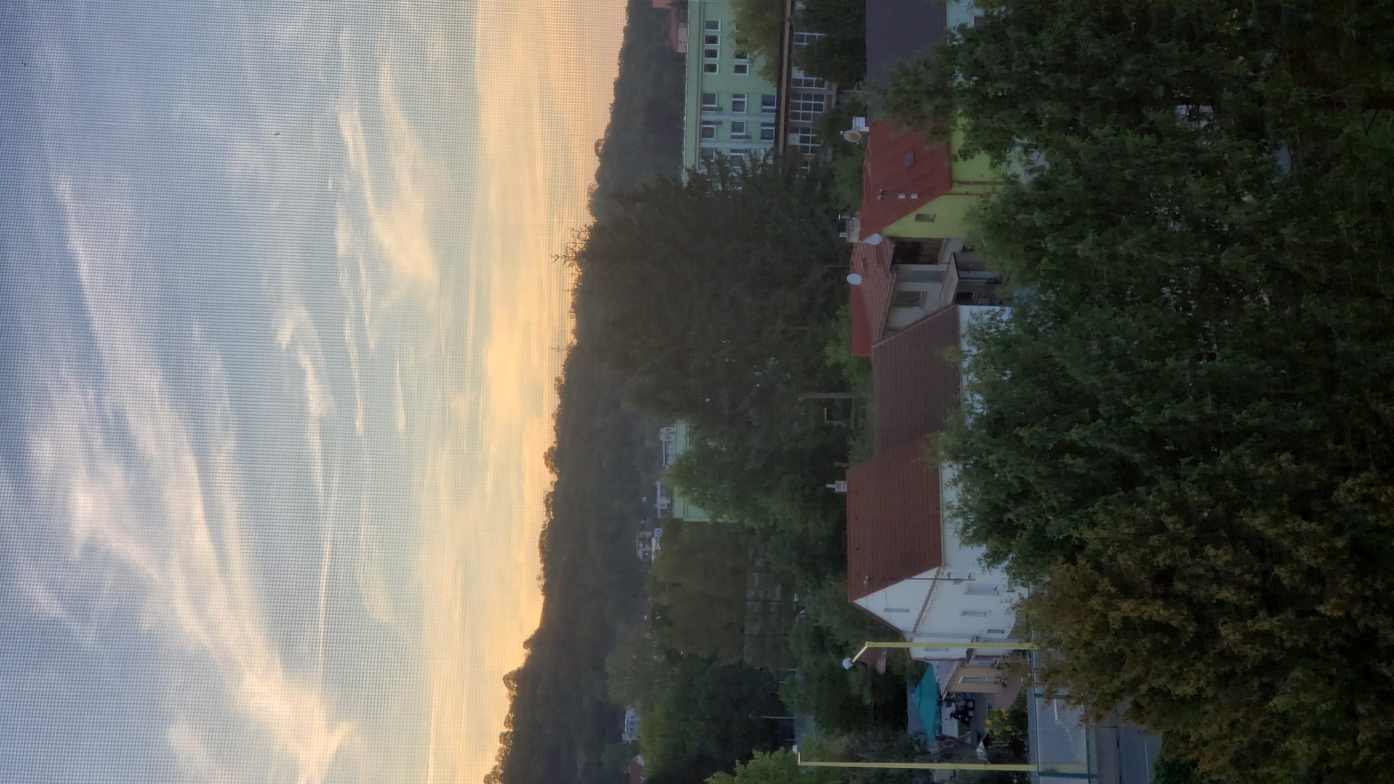 The image captures a view of residential buildings and a wooded hill at sunset. The scene appears to be from a higher vantage point, possibly an apartment building or a hill overlooking the neighborhood. In the foreground, several houses with red-tiled roofs are visible, nestled amongst lush green trees. Behind the houses, a larger, light green building with many windows stands out. The background is dominated by a tree-covered hill that slopes upwards, with the setting sun casting a warm, orange glow on the horizon and the sky above. The sky itself is a mix of blue and soft yellow, with wispy clouds streaking across it. The presence of a fine mesh pattern overlaid on the image suggests it was taken through a screen or window. The overall impression is a peaceful, suburban evening scene. This view is consistent with the landscape found in the Sekčov district of Prešov, Slovakia, where apartment blocks and residential areas are often situated near wooded hills.