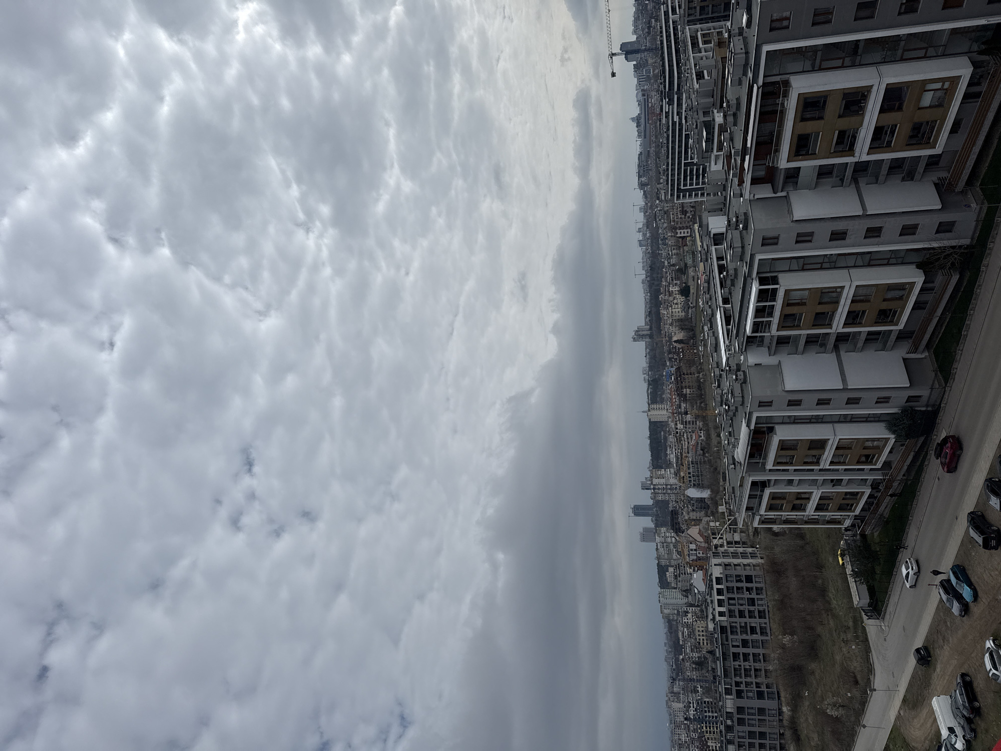 An aerial view of Sofia, Bulgaria under an overcast sky. Tall residential buildings in the foreground, followed by dense urban landscape stretching to the horizon. Several construction cranes visible among the buildings. Cars parked along the street in the bottom right. No people visible.