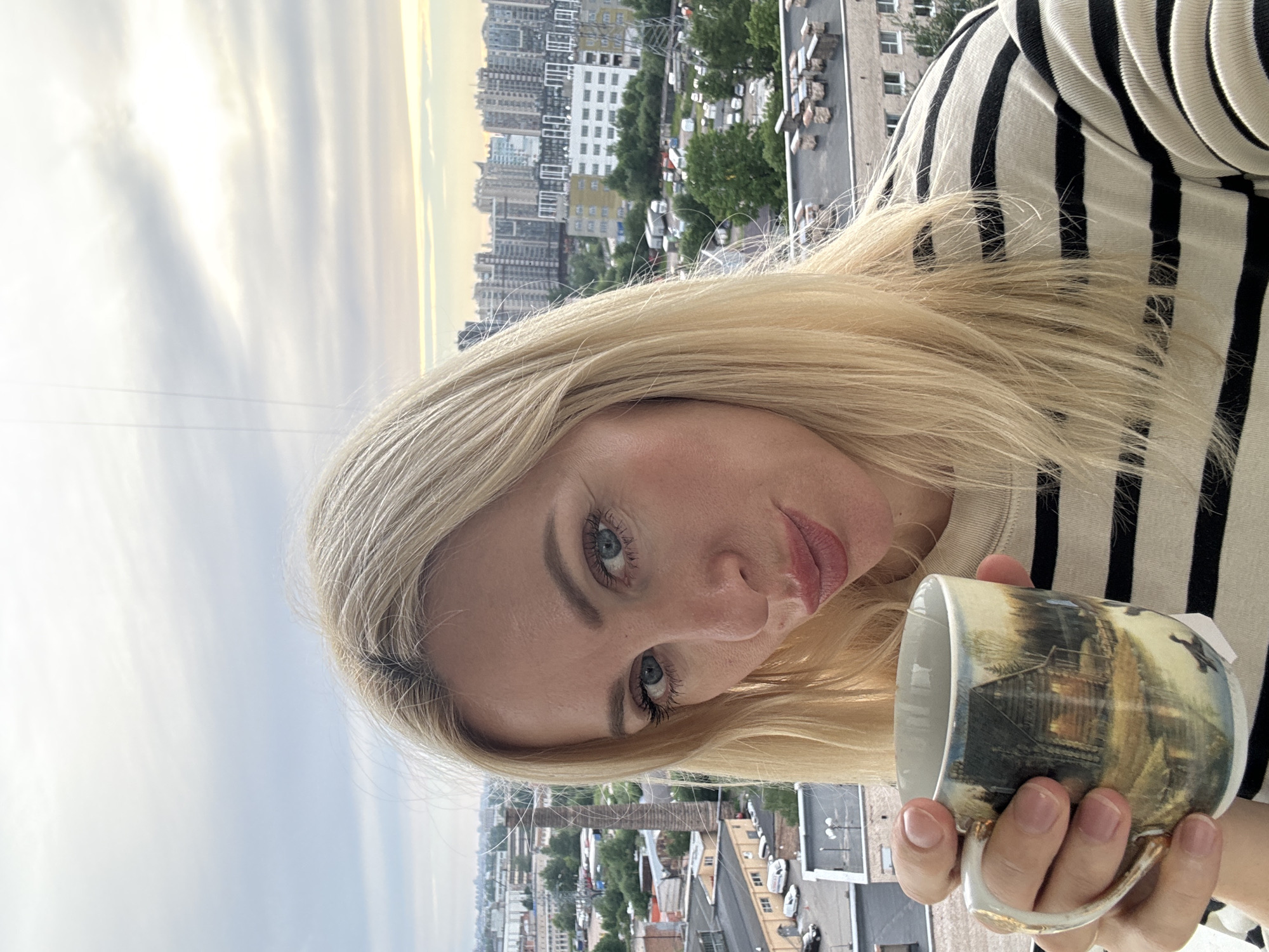 A woman with blonde hair is holding a mug with a landscape painting on it. She is wearing a striped shirt and looking at the camera. The background shows a city skyline with buildings and trees. The sky is cloudy.