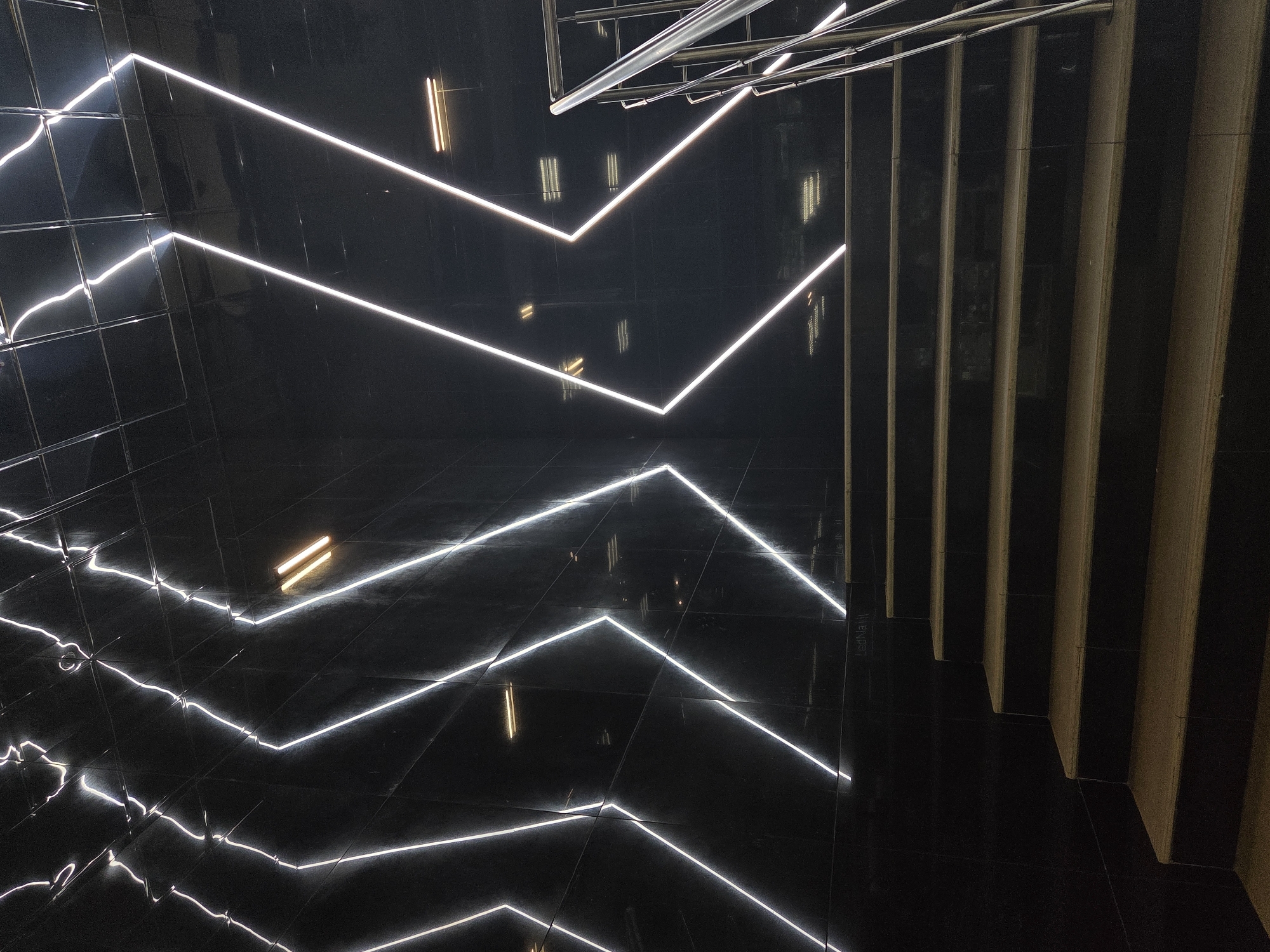 The image shows an interior space in an unknown city. The focus is on an area with glossy black tiles, possibly flooring or wall covering. Illuminated zig-zagging lines are prominent, reflecting on the tile surfaces. To the right are vertical panels, potentially wood or similar material, creating a textured wall or screen effect. A metal structure is visible at the top of the image.