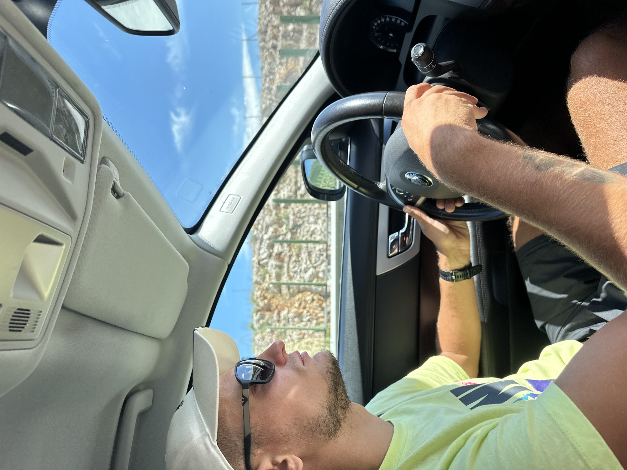 A man wearing a white baseball cap, sunglasses, and a neon yellow t-shirt is driving a Jaguar in Pelev Brijeg, Montenegro. The car's interior is visible, including the steering wheel, dashboard, and part of the ceiling. The man's hands are on the steering wheel. The exterior scenery shows a rocky hillside with sparse vegetation and a clear blue sky. The sun is shining brightly, casting shadows inside the car.