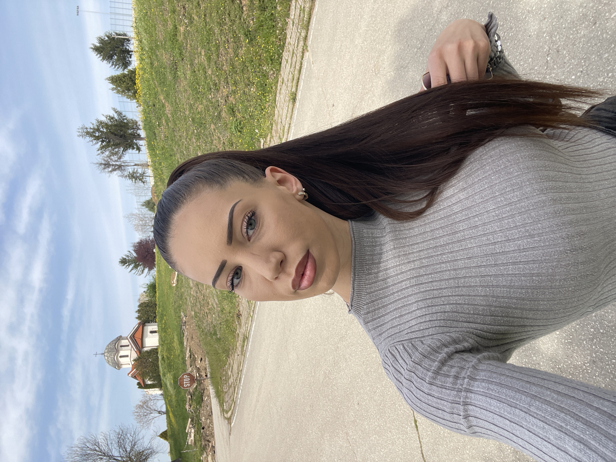 A woman with dark brown hair and light eyes stands on an asphalt road near a curb. She wears a gray ribbed top and has her hair pulled back. A white Orthodox church with an orange roof is in the background, along with trees and grass. A "STOP" sign is visible. The sky is partly cloudy. This scene appears to be in or near the city of Veliko Tarnovo, Bulgaria, judging by the church's architectural style.