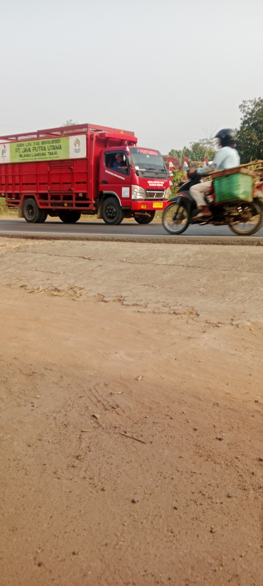A red delivery truck with "AGEN LPG 3 KG BERSUBSIDI PT. JAYA PUTRA UTAMA" written on its side is driving on a road. The truck has a load of what appears to be gas canisters covered by a green tarp. To the right of the truck, a person on a motorcycle is also driving. The person is wearing a blue shirt and a helmet. The motorcycle has a green basket attached to the back. The scene takes place on a paved road with dry, cracked dirt and sparse vegetation on the sides. The sky is overcast and a few trees are visible in the background. The image appears to have been taken in Gondang Rejo, Indonesia.
