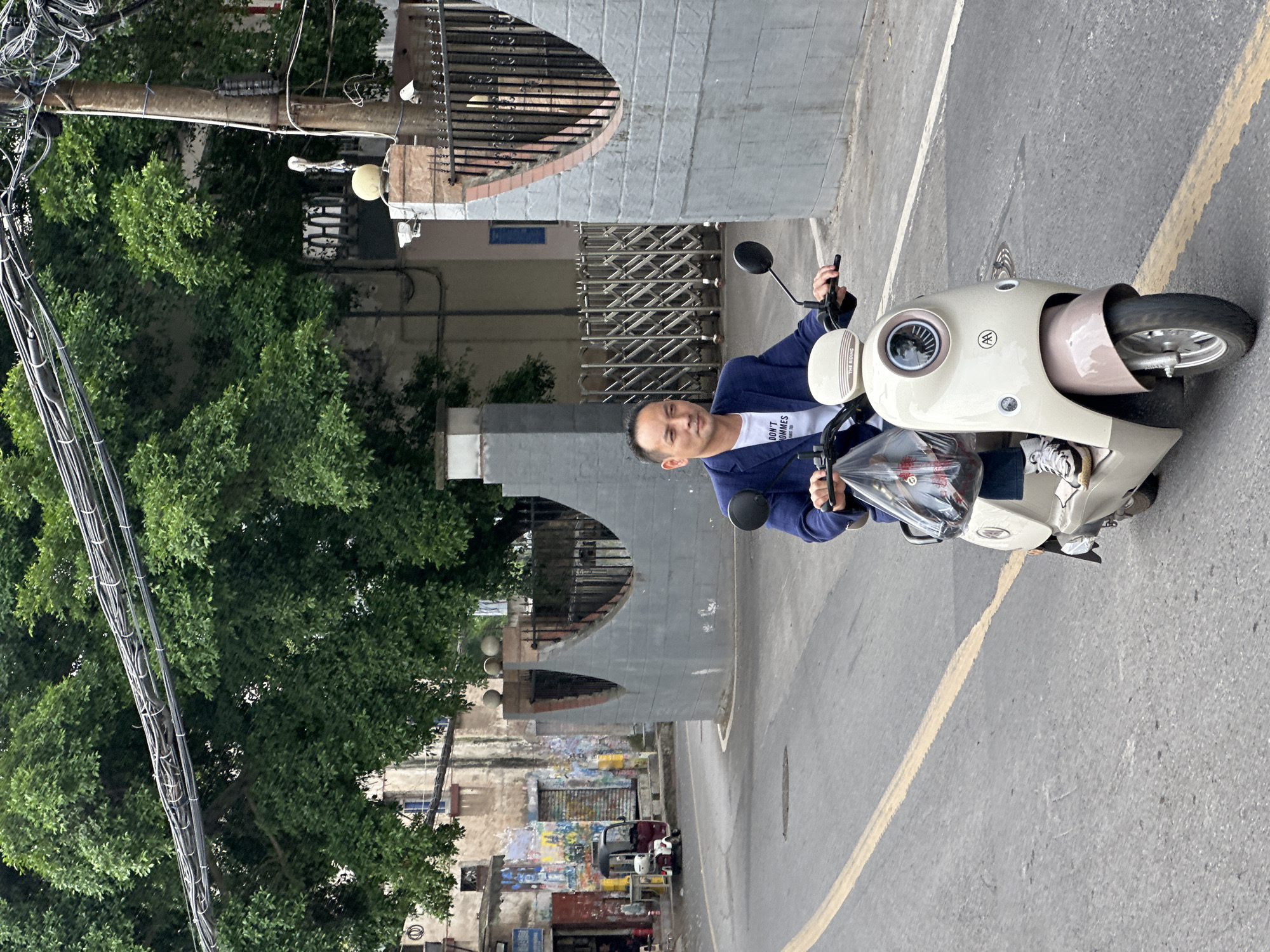 A man is sitting on a moped in a street scene. The man is wearing a blue blazer over a white t-shirt and white sneakers. The moped is light beige with brown accents. The street is paved with a yellow line marking traffic lanes. Buildings are visible in the background, including a gray concrete structure with arched openings. A tree with dense green foliage is on the left side of the frame. Overhead power lines are also visible. The scene is located in the Jiulongpo District, China.