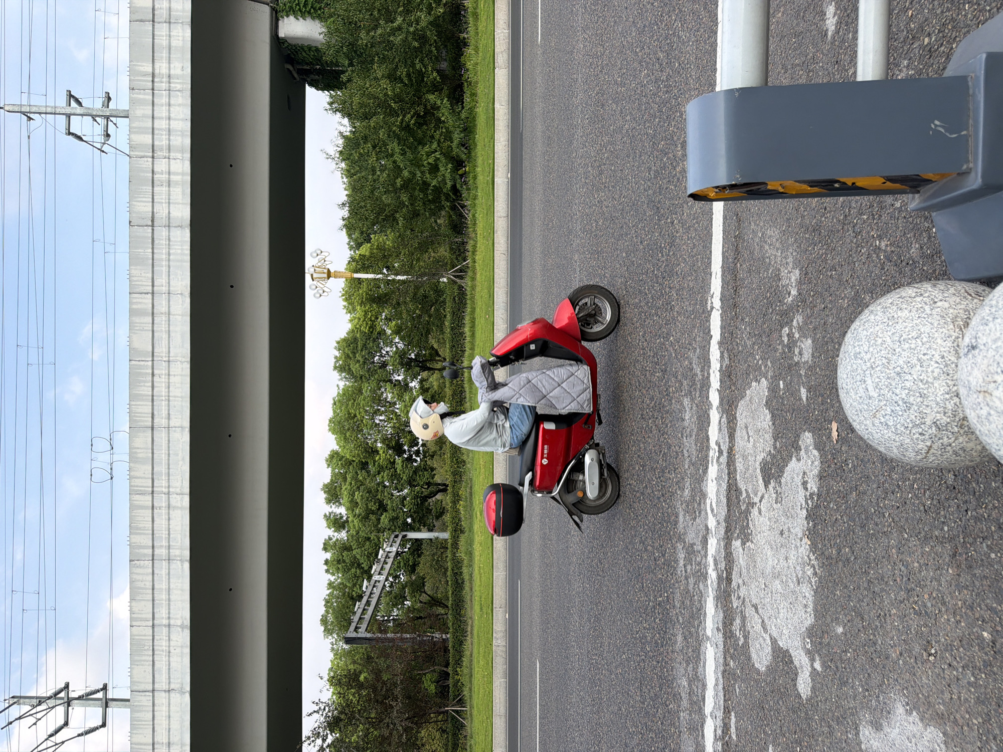 A person wearing a light-colored long-sleeved shirt, blue jeans, a light-colored helmet, and a face mask rides a red electric scooter on a paved road in Jiangjin, China. The scooter has a black storage box on the back and a quilted gray cover over the rider's legs. The road has white lane markings and some water or oil stains on the surface. In the background, a large concrete overpass runs horizontally across the image. Beyond the overpass, lush green trees line the road, and power lines are visible against a cloudy sky. To the right of the scooter, a gray and yellow traffic barrier is partially visible. The overall scene appears to be an urban or suburban street environment.