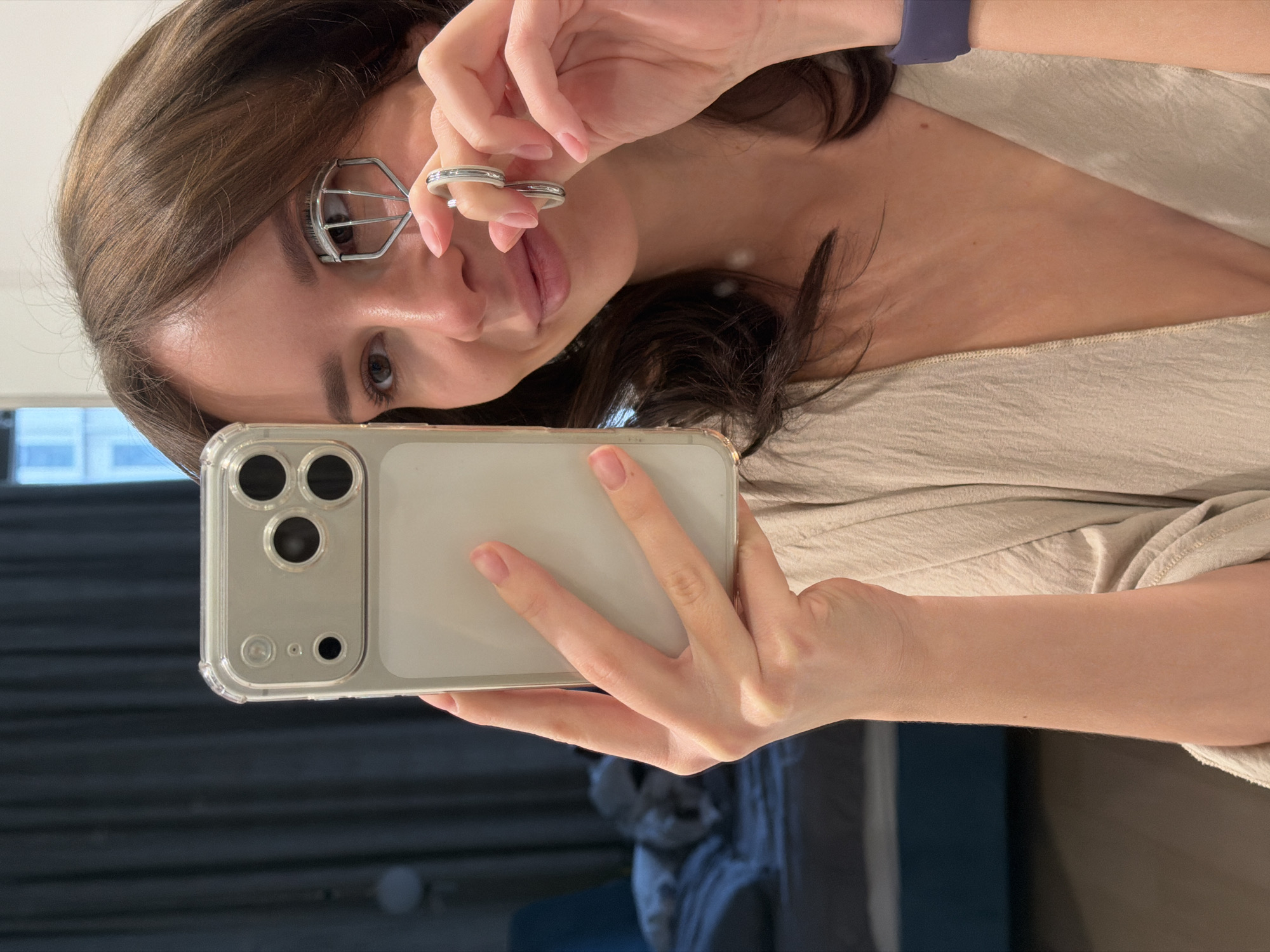 A woman is holding a smartphone with a clear case in her left hand, taking a selfie in what appears to be an indoor setting. She is using an eyelash curler on her left eye. The backdrop suggests a modern interior with a glimpse of what could be an urban landscape through a window. The image's location is not discernible from the image alone; therefore, stating the location is in Moscow, Russia, is not possible based on visual evidence.