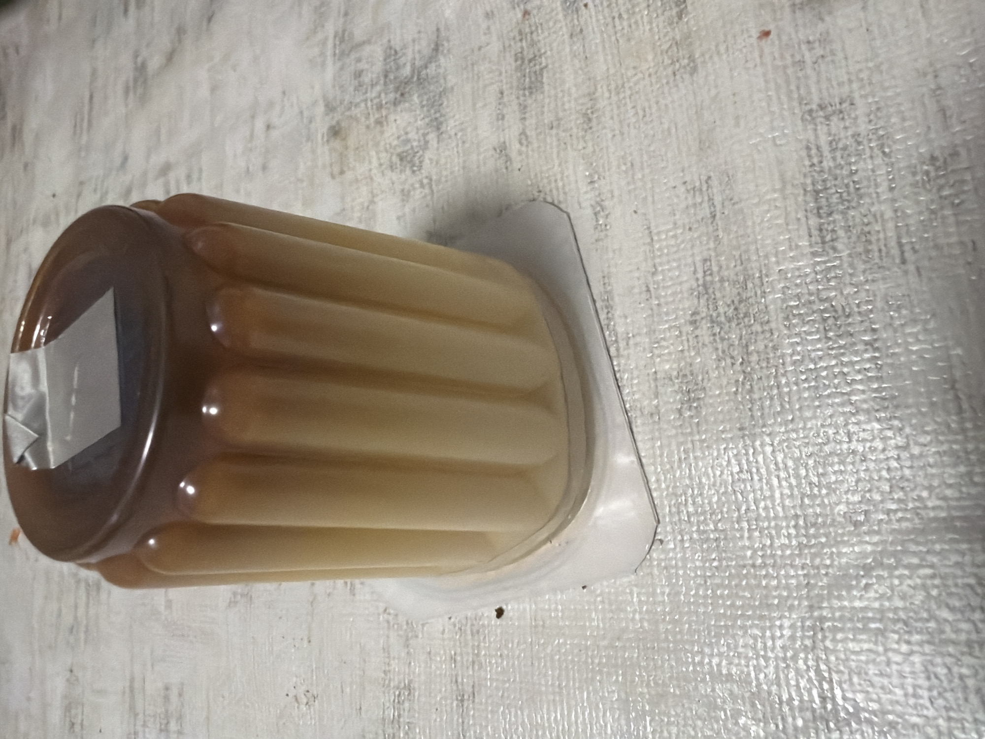 A close-up, eye-level shot shows a fluted pudding in a clear plastic container. The pudding is a gradient of pale yellow to a rich brown caramel color at the top, with the caramel layer forming a smooth, glossy surface. The plastic container is square with rounded corners and a slightly raised rim. The pudding rests on a textured, white surface that appears to be a tablecloth or placemat. The lighting is overhead, creating subtle highlights on the pudding's surface and casting a soft shadow. There are no people or other objects clearly visible in the frame.