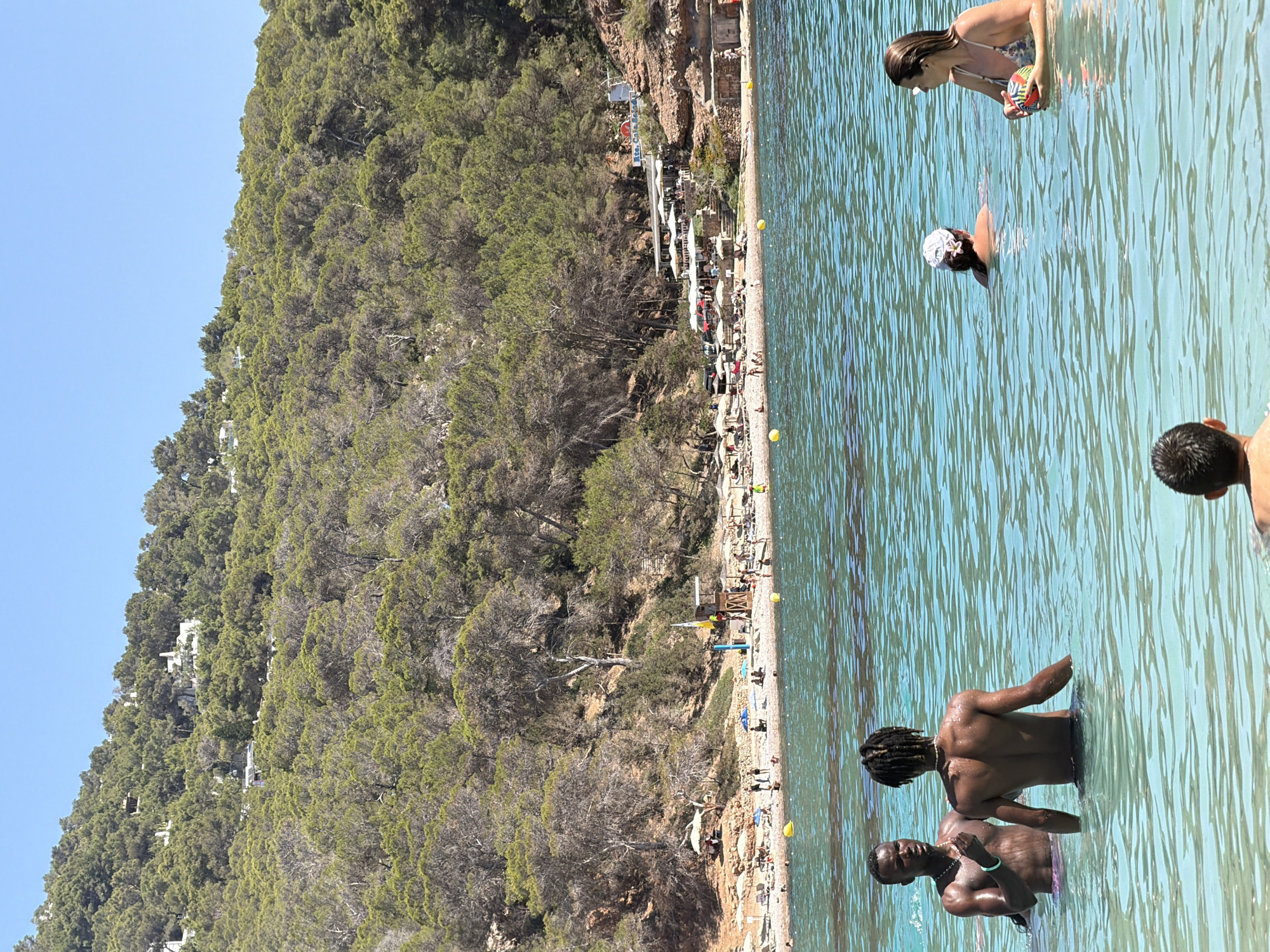 The media file displays a vibrant, sun-drenched beach scene at Sant Antoni de Portmany, Spain. The foreground is dominated by clear, turquoise water with gentle ripples reflecting the bright sunlight, suggesting a warm, pleasant day. Several people are visible in the water. In the middle ground, a group of men stand in the water, their torsos exposed to the sun. Further back, a woman in a bikini is holding a colorful ball, and another person with a white cap is submerged in the water.
The background features a rocky coastline densely covered with lush green trees and vegetation. White buildings are nestled amongst the trees, indicating a developed area bordering the natural landscape. Along the shoreline, a beach is lined with umbrellas and lounge chairs, suggesting a popular tourist destination. A small establishment with tables and umbrellas is visible at the base of the cliff. Yellow buoys are scattered in the water. The overall atmosphere is one of relaxation and enjoyment, typical of a summer vacation day at the beach. The clear blue sky indicates fair weather.