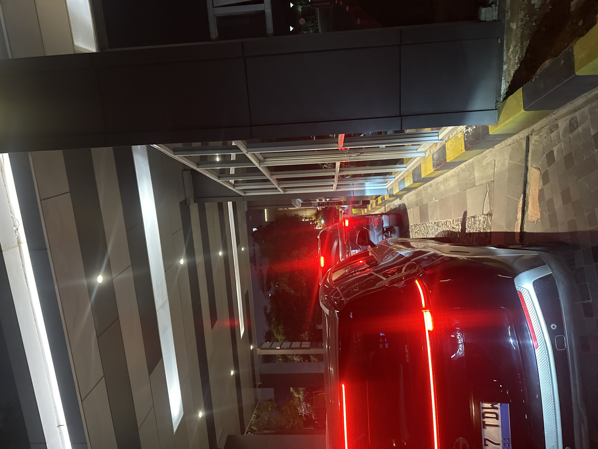 The image depicts the rear of a dark-colored car, its tail lights brightly illuminated. The car is positioned on a street or driveway, flanked by a building on the left with a modern facade, featuring alternating light and dark vertical panels. The right side reveals a curb with alternating yellow and black hazard markings, transitioning into a rough pavement or ground surface. The location is likely Central Jakarta, Indonesia, indicated by the vehicle's license plate. Other cars are visible in front of the main car, creating a sense of a queue or line of vehicles. The scene appears to be at night.
