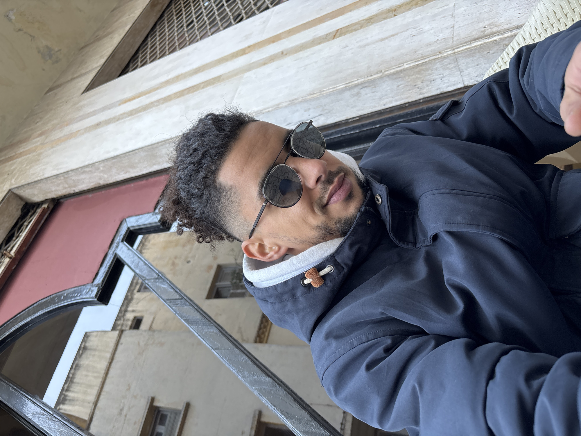 A man with dark curly hair, a beard, and sunglasses is pictured from a low angle in what appears to be Tanger, Morocco. He is wearing a dark blue jacket with a white-hooded liner. The background shows a building with ornate architectural details and windows. The image is taken outdoors on a balcony or terrace, as suggested by the wrought iron railing in the foreground.