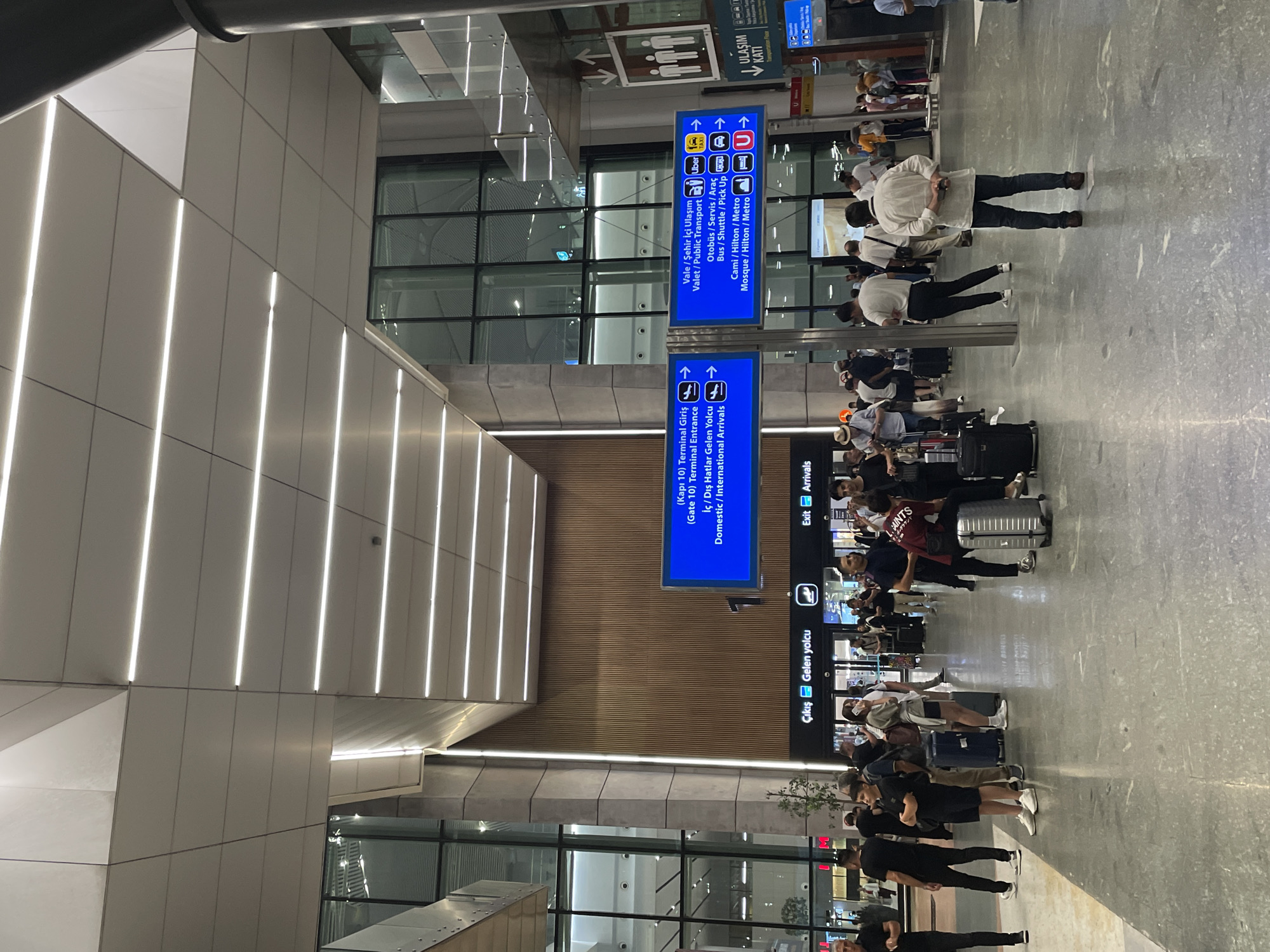 The image shows a busy interior of an airport terminal in Arnavutköy, Turkey, likely Istanbul Airport given the bilingual signage. The architecture features a modern design with a high ceiling illuminated by linear lights. Various travelers are seen in the foreground and midground, some standing with luggage, others walking.
In the midground, there are several large blue digital signs displaying directional information. One sign indicates "(Kapı 10) Terminal Giriş / İç / Dış Hatlar Gelen Yolcu / Domestic / International Arrivals" with arrows pointing right. Another sign lists destinations like "Vale / Şehir İçi Ulaşım / Valet / Public Transport," "Otobüs / Servis / Araç / Bus / Shuttle / Pick Up," and "Cami / Hilton / Metro / Mosque / Hilton / Metro," also with directional arrows to the right, accompanied by icons for Uber, taxi, bus, and metro.
Below these signs, a horizontal panel displays "Çıkış | Gelen Yolcu" (Exit | Arrivals) with a plane icon, and "Exit Arrivals" with a corresponding arrow.
People are dressed in casual attire. Many have wheeled luggage, suggesting travel. The flooring is a polished grey material. In the background, large glass windows reveal more of the airport's structure, including escalators and possibly other terminals. There is also signage for "ULASIM KATI" (Transportation Floor). The overall atmosphere appears to be one of transit and movement.