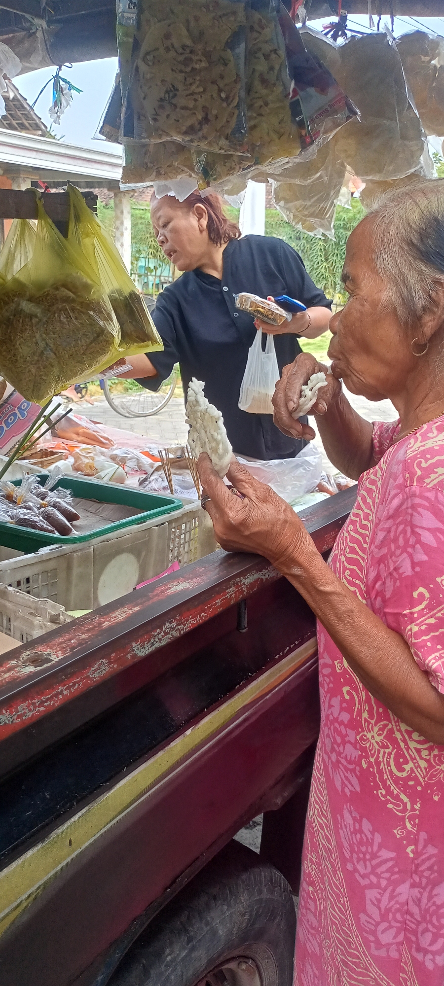 An elderly woman in a pink patterned dress is standing at a market stall in Gempolklutuk, Indonesia, and eating a white, puffed snack. Her right hand holds the snack up to her mouth, while her left hand is holding another piece of the snack. To her left, a woman wearing a black shirt appears to be attending the stall, holding plastic bags containing snacks. The stall is stocked with various packaged snacks, some in yellow plastic bags, and others in clear plastic packaging. Several green and white plastic containers are also visible, holding more packaged snacks. The stall itself is constructed of weathered wood, painted in shades of red and yellow. The ground beneath the stall is visible, with a tire of a vehicle peeking out from the lower edge of the frame. The background suggests an outdoor setting, with greenery and some buildings visible.