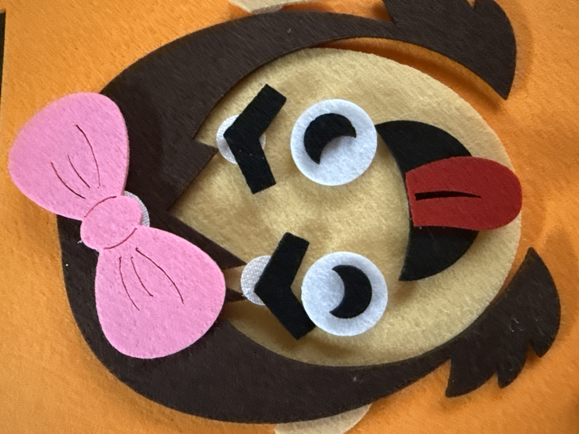 A close-up shows a felt owl craft project. The owl has an orange background, a brown outer face, a tan inner face, white eyes with black crescent pupils, black felt eyebrows, and a red felt beak. A pink felt bow is attached to the left side of the owl's head. The image is set indoors under bright lighting.