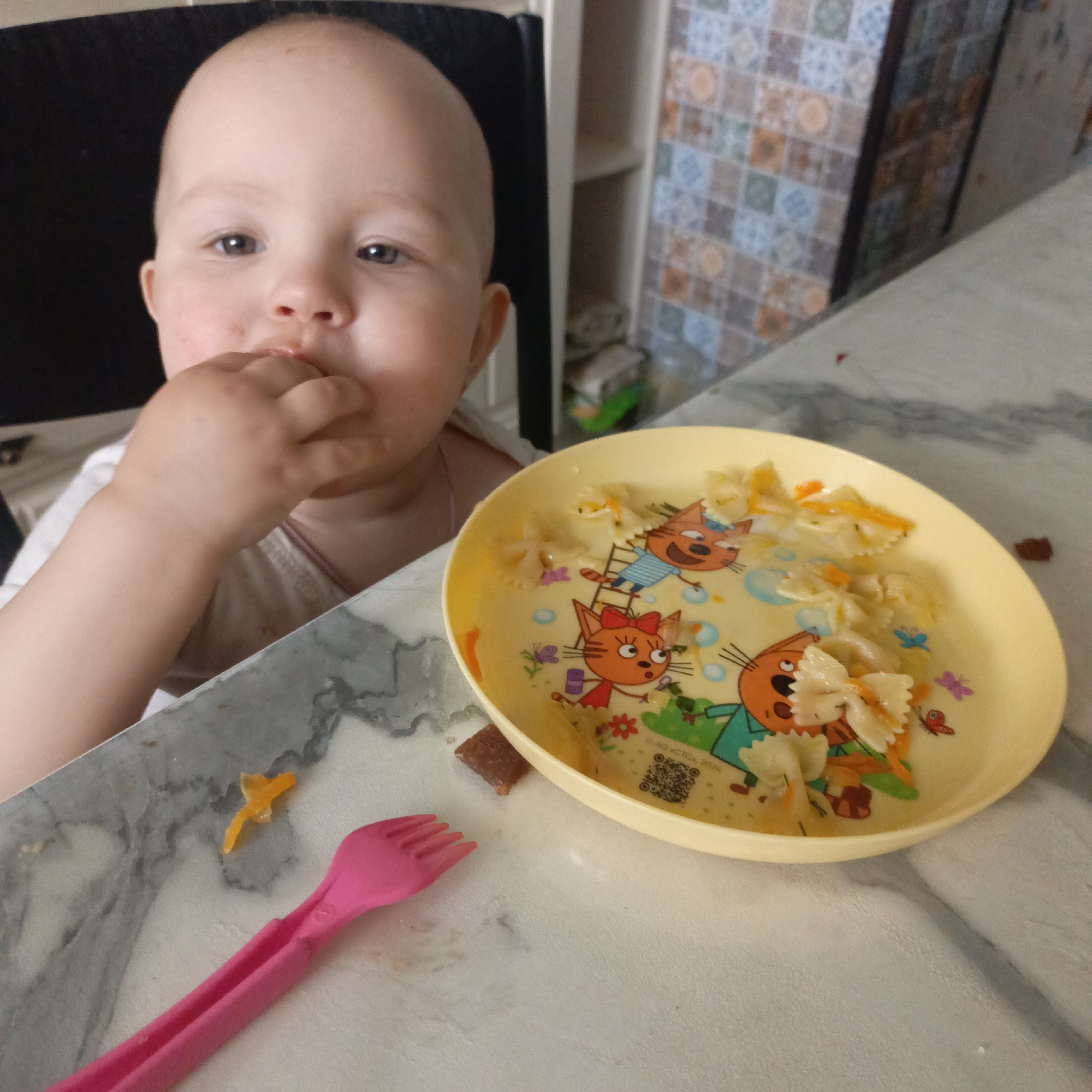 A young child or toddler is pictured in a close-up, eating at a table. The child, with fair skin, dark eyes, and a bald head, is positioned on the left side of the frame, with their left hand near their mouth, suggesting they are bringing food to their mouth or have just done so. They are wearing a light-colored top.
On the table in front of the child, to the right, is a yellow plastic bowl decorated with cartoon cats, butterflies, and flowers. The bowl contains farfalle (butterfly-shaped pasta) mixed with small orange pieces, possibly carrots. A pink plastic fork lies on the table to the left of the bowl. The table surface is light-colored, with a marble-like pattern. Several small food remnants are scattered on the table, including an orange piece near the fork and a dark crumb near the bowl.
The background features a tiled wall with a repetitive geometric pattern in shades of blue, orange, brown, and white. To the left of the tiled wall, a light-colored shelf or cabinet is partially visible. A dark, possibly black, object is seen behind the child's head, likely the back of a chair.
The setting appears to be an indoor mealtime, likely in a kitchen or dining area. The child's expression is neutral, and they are looking slightly to their right. Visible text on the bowl includes a QR code and copyright information: "© AO “CTCL” 2024".