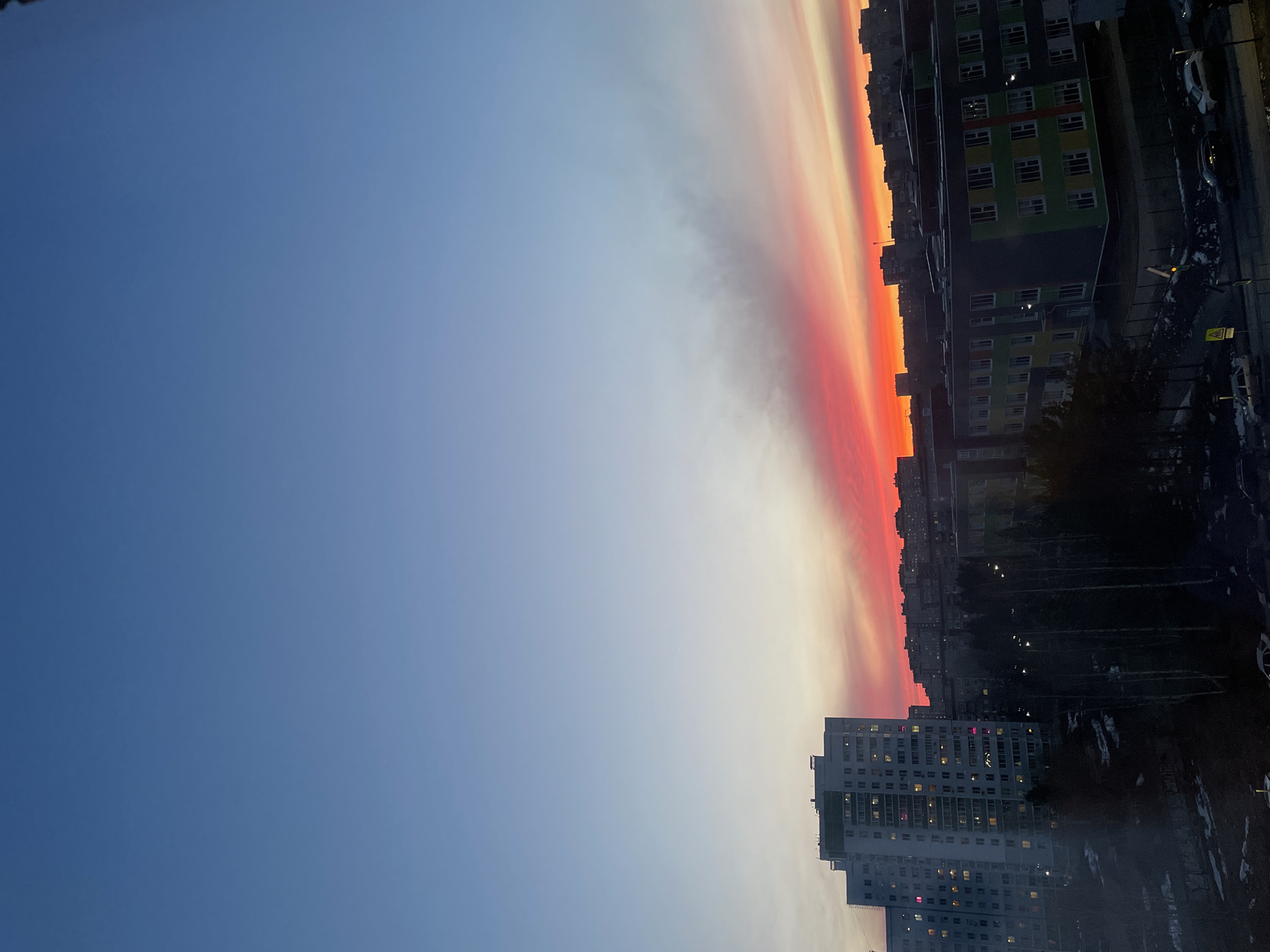 The image shows a sunset over a city, specifically Izhevsk, Russia. The sky transitions from a deep blue in the upper left to a band of orange, red, and white at the horizon. Silhouettes of buildings line the lower right side of the image, including a large apartment building with many illuminated windows. A road with cars is visible on the right. Some trees are also present. The view is from a high vantage point, possibly from another building.