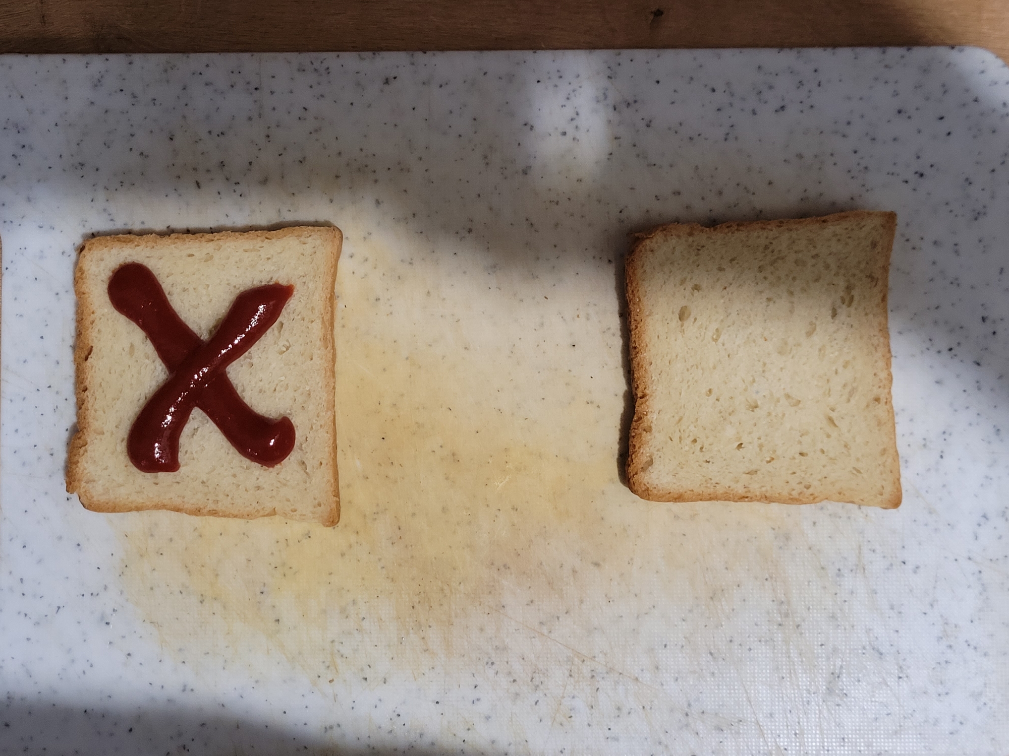 On a speckled white cutting board, two slices of white bread are laid out. The top slice has a red 'X' shape drawn on it with what appears to be ketchup. The bottom slice of bread is plain. The cutting board is situated on a wooden surface. There is no indication of a city or location.