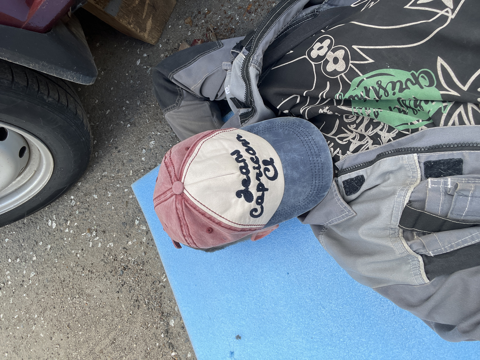 The image shows a close-up, overhead view of a person lying on a blue foam mat on asphalt. The person is wearing work clothes, including gray overalls with white stitching and a black shirt with white line drawings and a green graphic. A red and blue baseball cap with white lettering is on the person's head. The cap has the words "Jean" and "Caprice" embroidered on it in a dark color. In the background, a car tire and part of a car's fender are visible, as well as a wooden object. The lighting suggests it is daytime, likely outdoors, possibly in a workshop or garage setting. There are no clear indicators of the weather or specific emotions. The location context provided is Veteran-2, Russia.