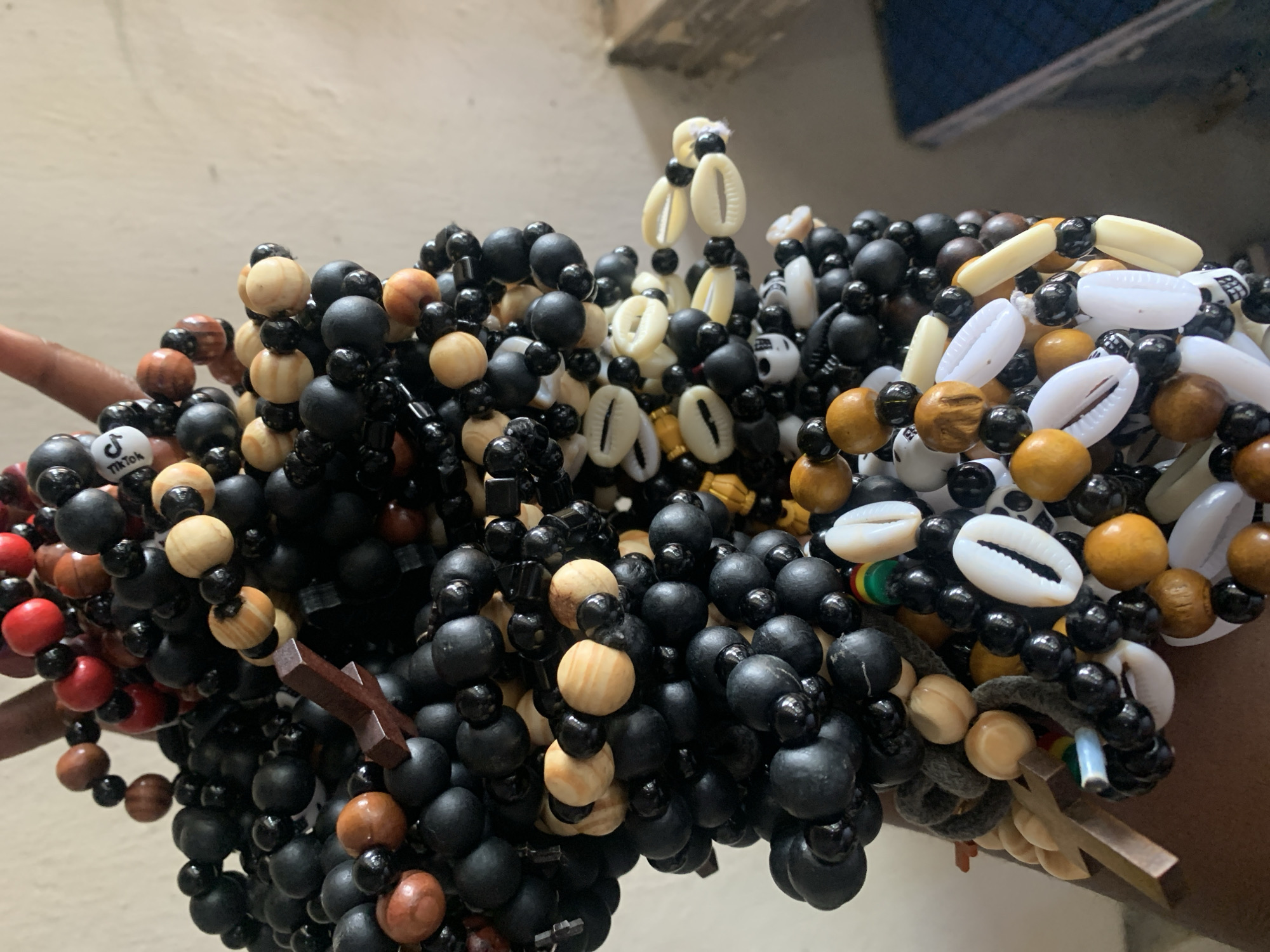 The image shows several strands of beaded bracelets held in a person's hand. The bracelets are made of beads in various colors, including black, brown, red, and white. Some of the bracelets also incorporate cowrie shells. One bracelet appears to have a small wooden cross attached to it. Another has a bead with the TikTok logo on it. The location, Kabong, Nigeria, is not visually discernible from the image.