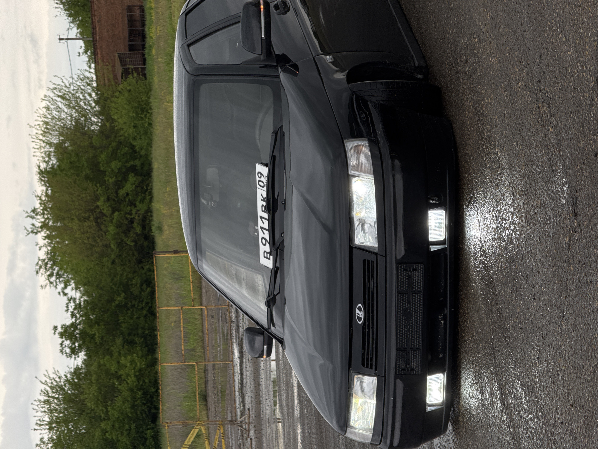 The image shows the front of a black car, specifically a Lada or similar model, with its license plate visible. The license plate reads "B911PK09". The car is parked on wet asphalt. In the background, there's a blurred view of green foliage, possibly trees and shrubs, along with what appears to be a rusty metal structure or building. The sky is overcast. Considering the "09" on the license plate, the car may be from the Republic of Kalmykia, Russia. While a specific location cannot be definitively confirmed to be Pavlovskaya, Russia from the image alone, the scene suggests a rainy day in a slightly industrial or semi-rural area within Russia, given the car model, license plate, and background elements. The car lights are on. There is an orange structure in the background.