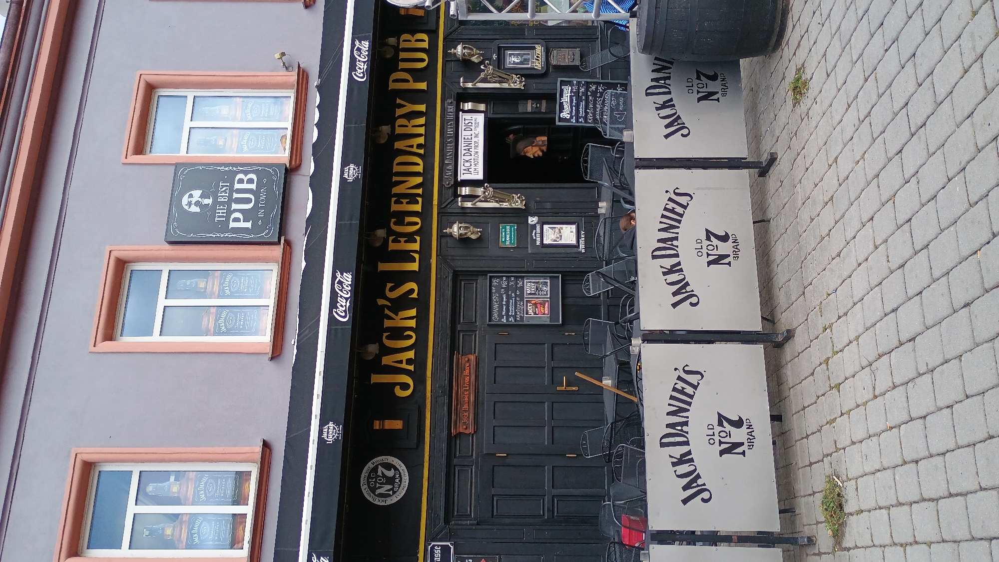 The image depicts the exterior of "Jack's Legendary Pub" located in Sokolov, Czechia. The pub's facade is dark, featuring a large sign with the pub's name in yellow lettering. Above the entrance, there are two windows decorated with images of Jack Daniel's bottles. A prominent black awning stretches across the building, adorned with Coca-Cola logos on both ends and "Jack's Legendary" in the center. To the right of the pub entrance, a sign reads "THE BEST PUB IN TOWN."
In front of the pub, there are tables and chairs set up on a cobblestone patio, suggesting an outdoor seating area. White panels with "Jack Daniel's Old No. 7 Brand" logos are arranged as barriers on the patio. A dark barrel is visible to the right of these panels. The building itself has a light gray exterior with salmon-colored window frames. The overall scene appears to be during daylight.