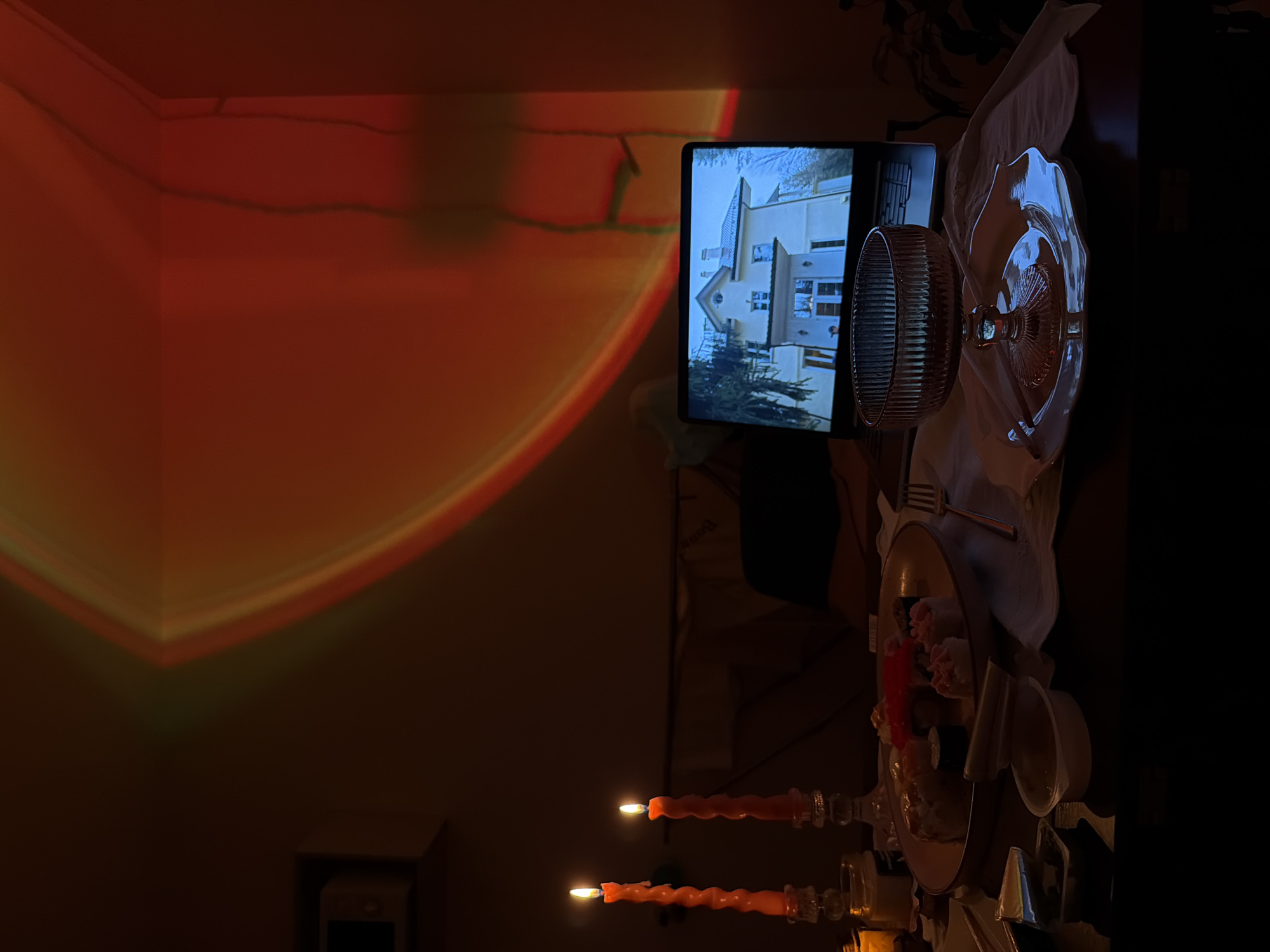 The scene is a dimly lit interior. Light emanating from two lit, orange, candle-shaped objects illuminates the base of a table, which has a dinner setting on top. The setting includes clear plates, bowls, silverware, and a light-colored cloth. A monitor is turned towards the table, and displays a picture of the exterior of a light-colored house surrounded by green trees. Light in red and orange hues are reflected in a corner of the room. The specific location in Izhevsk, Russia cannot be ascertained from this image alone.