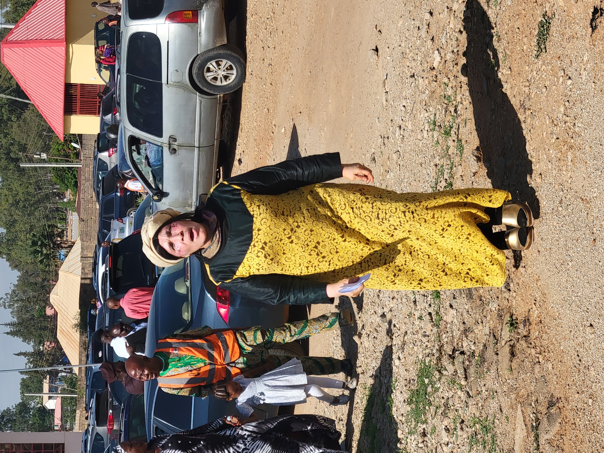 The image depicts a woman in a yellow lace dress standing in what appears to be a parking area in Gura Top, Nigeria. A row of parked cars is visible in the background. Other people are present in the vicinity, including a man in an orange vest and a child in a white dress. The ground is mostly dirt and gravel. A building with a red roof can be seen in the background.