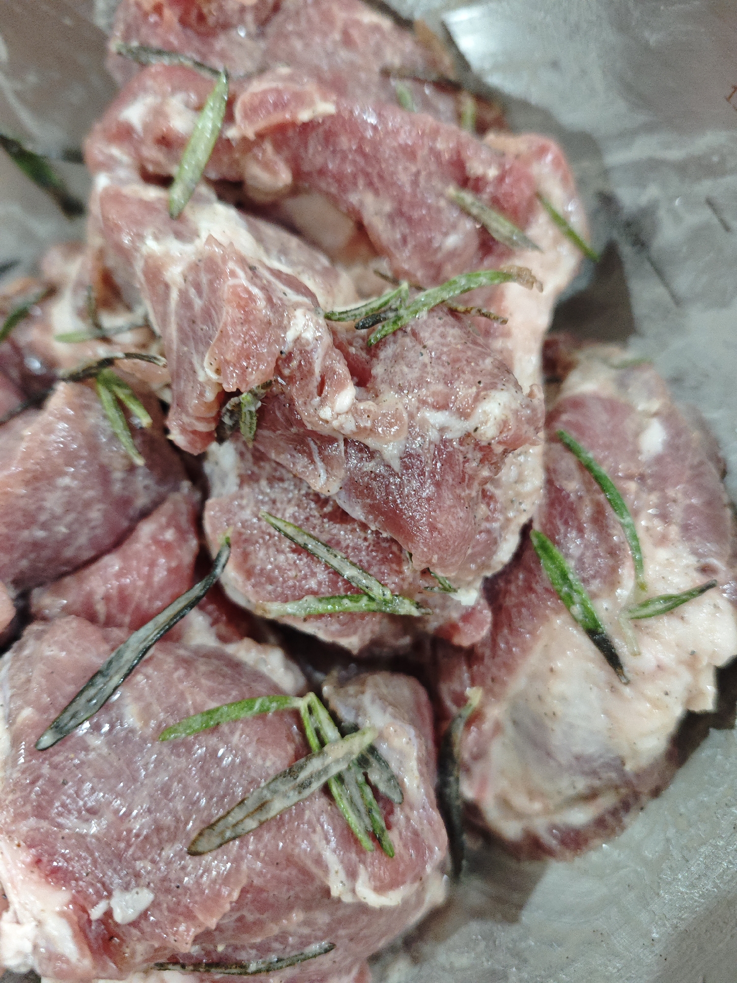 The image shows raw, cubed pieces of meat, possibly pork or lamb, seasoned with what appears to be fresh rosemary. The meat is pink with visible fat marbling. The scene is a close-up, likely in a domestic kitchen setting, inside a bowl. Location is unknown.