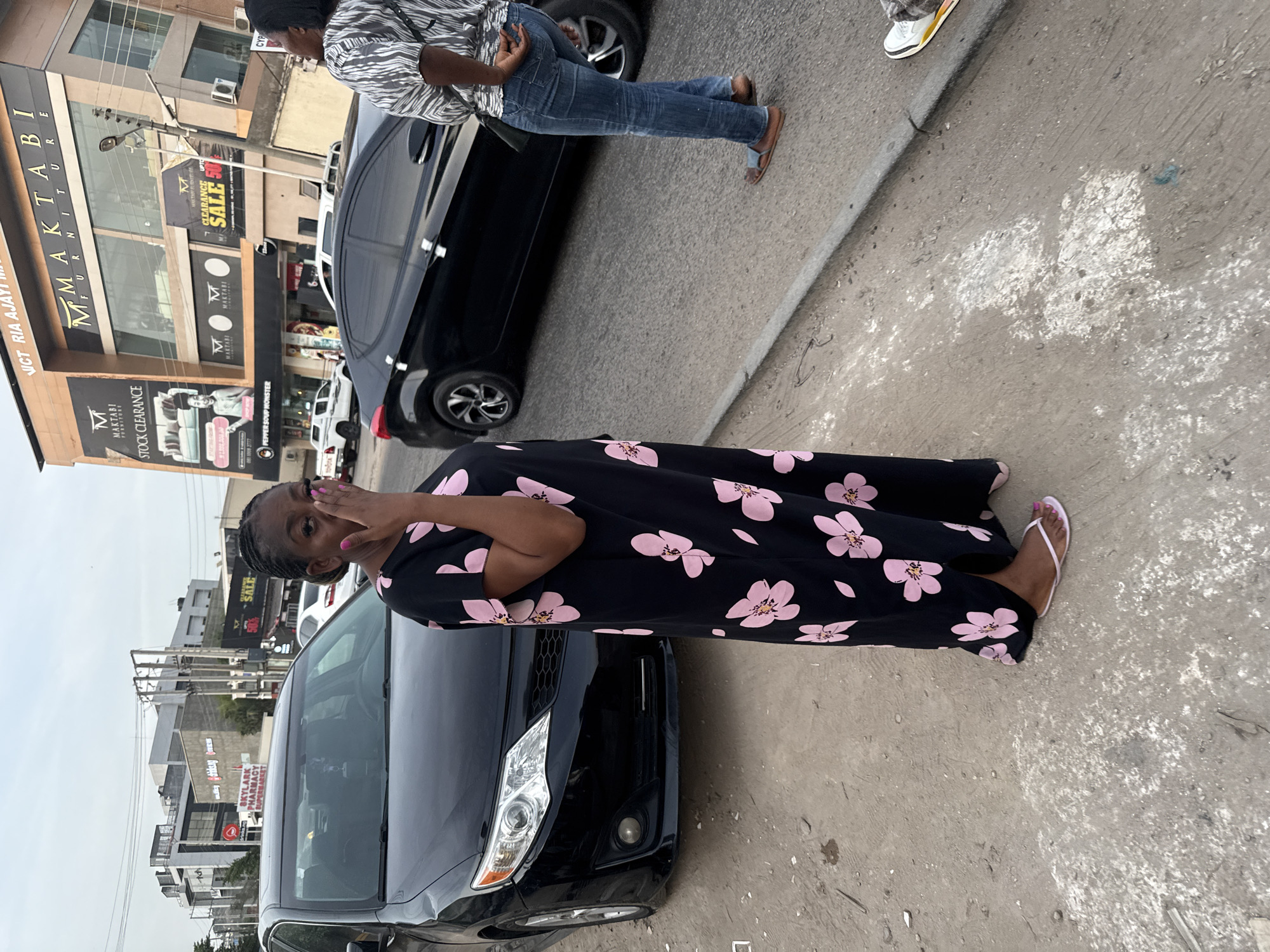 The image depicts a woman standing next to a black car on a city street in Gwarinpa, Nigeria. The woman is wearing a black dress with pink floral patterns. A building with the name "MMAKTABI FURNITURE" can be seen in the background, along with a sign mentioning "STOCK CLEARANCE" and "SALE". Other shops and street scenes are visible in the background. Another person is standing next to a black car nearby. The environment appears to be a commercial area with various buildings and traffic.