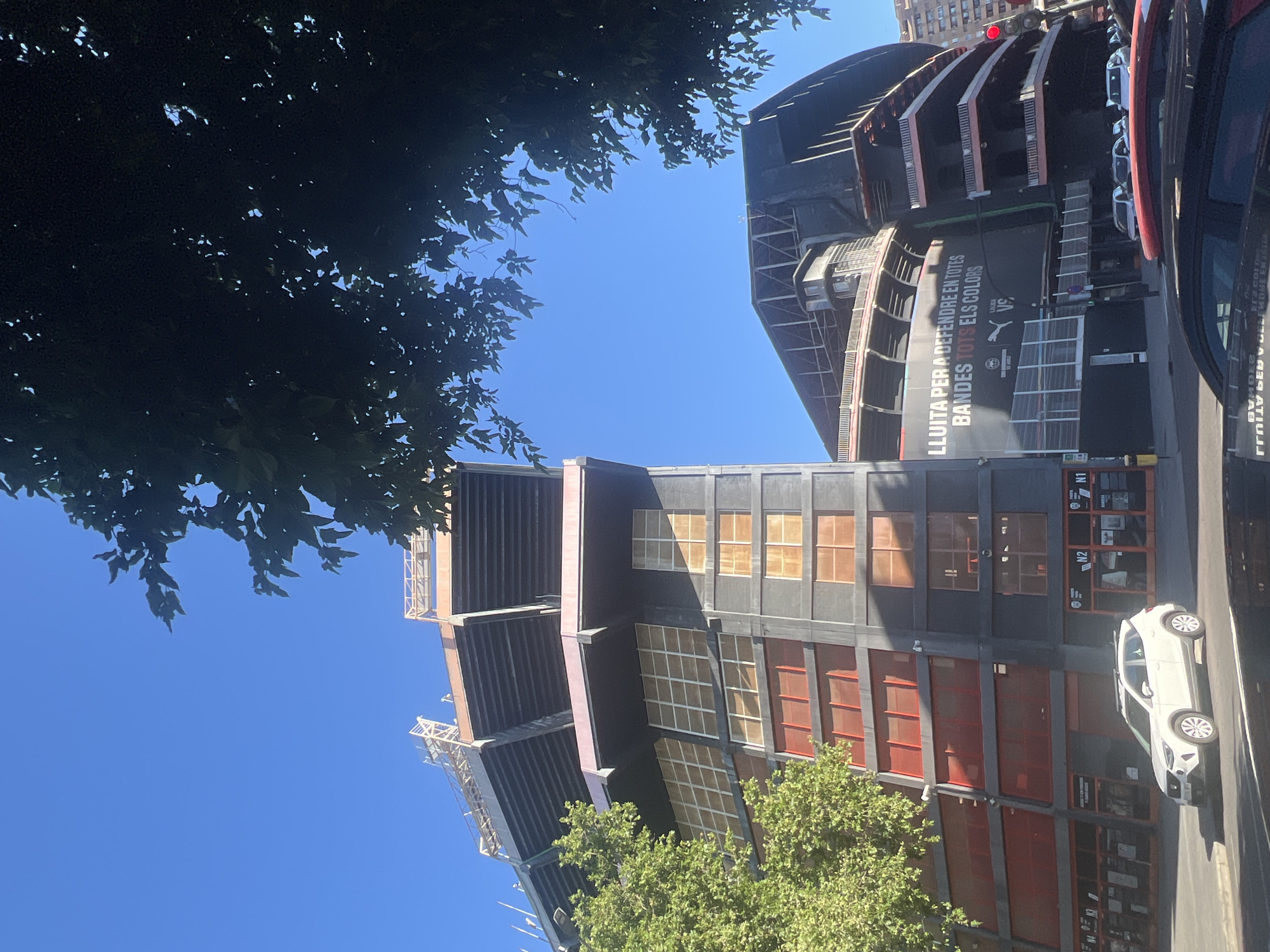 The image shows a partial view of the Mestalla Stadium in Valencia, Spain. The stadium's gray facade with red and orange window sections is visible. A large banner with the text "LLUITA PER A DEFENDRE EN TOTES BANDES TOTS ELS COLORS" is displayed. A white car is parked on the street in front of the stadium. Trees are visible on both sides of the stadium. The sky is blue.