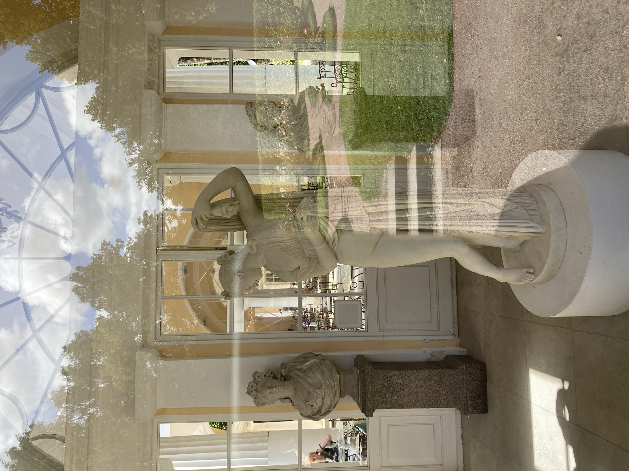 The image depicts a reflection on a windowpane, showing an interior and exterior scene. Inside, there is a white marble statue of a woman with her arm raised, draped in classical attire, standing on a circular pedestal. To the left, another white marble bust of a woman adorned with a floral wreath is displayed on a granite pedestal. A person is visible through the window, seated at a table.
The reflection also reveals the exterior, which includes a garden with green grass, a gravel path, and rose bushes. There are outdoor tables and chairs visible in the garden. The sky with clouds and trees is reflected on the upper part of the window. The architectural style of the building suggests it is located in a park or a palace setting. This scene is consistent with the Pavlovsk Park, an imperial park in the city of Pavlovsk, Russia, known for its neoclassical architecture and sculptures.