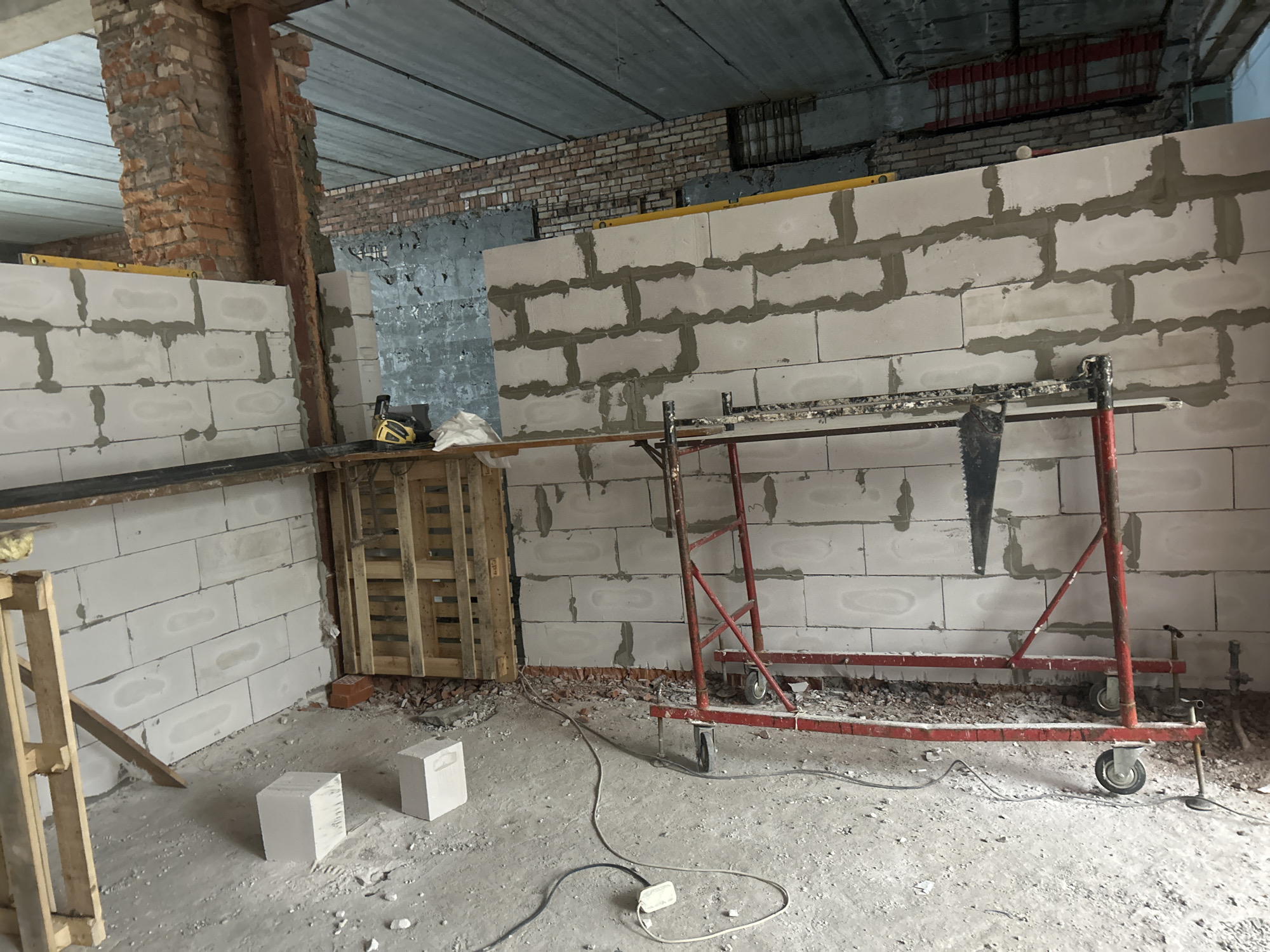 The image depicts an interior construction site in Saint Petersburg, Russia. The walls are constructed from white concrete blocks with visible mortar. A metal scaffolding structure with red paint and wheels stands in the center of the image, supporting a saw and other tools. To the left, a wooden pallet is leaning against a wall. In the foreground, two white concrete blocks rest on the dusty concrete floor, and electrical cords are scattered across the ground. The ceiling appears to be made of concrete slabs. The overall scene suggests ongoing construction or renovation work.
