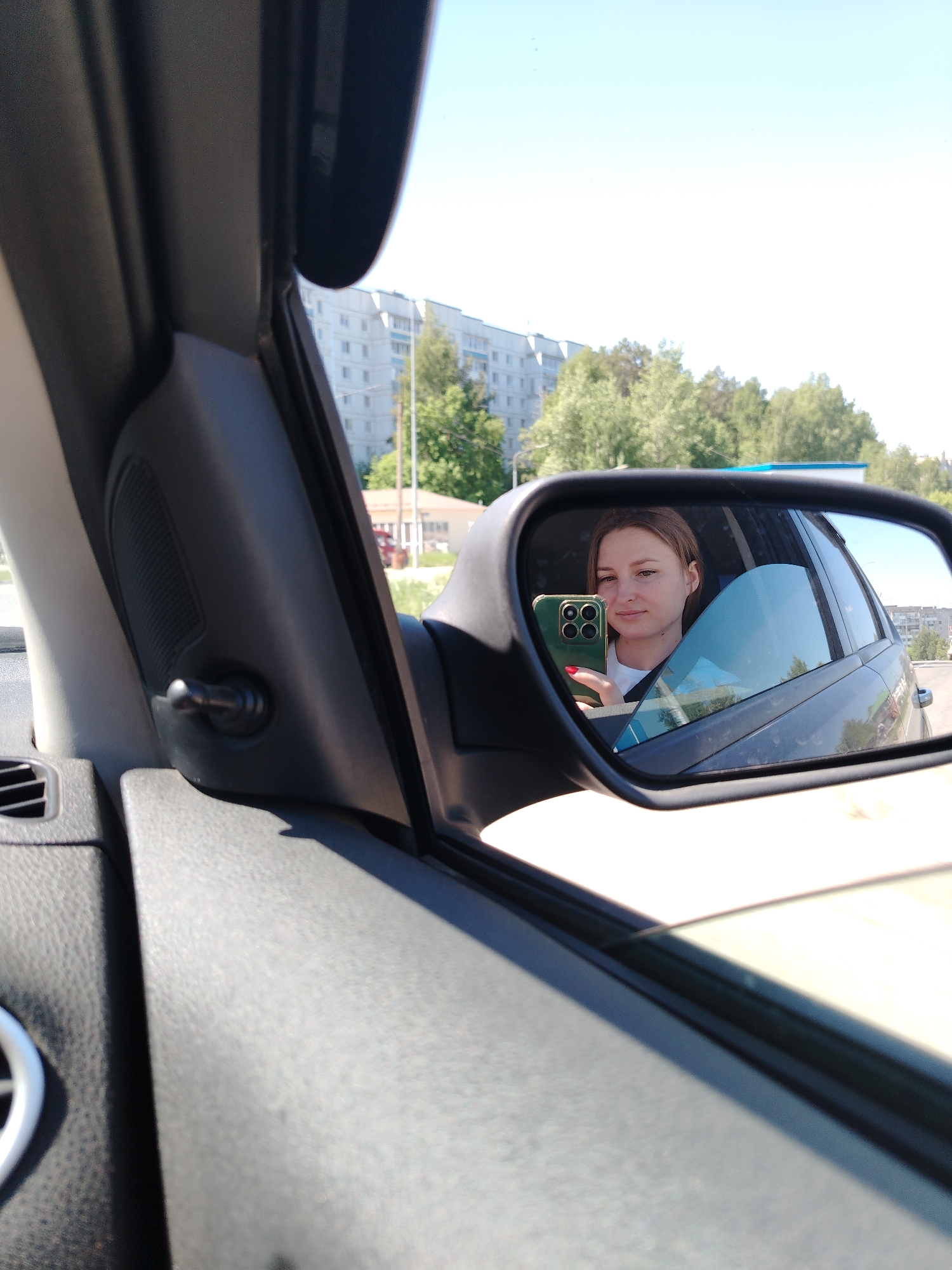 A woman with light brown hair is visible in the side mirror of a car. She is holding a green phone with multiple camera lenses and red nail polish is visible on her fingernails. Outside the car, a multi-story building and trees are seen. The interior of the car, including the dashboard and the side mirror casing, are visible. The location appears to be Chaikovsky, Russia based on the architectural style of the buildings in the background.